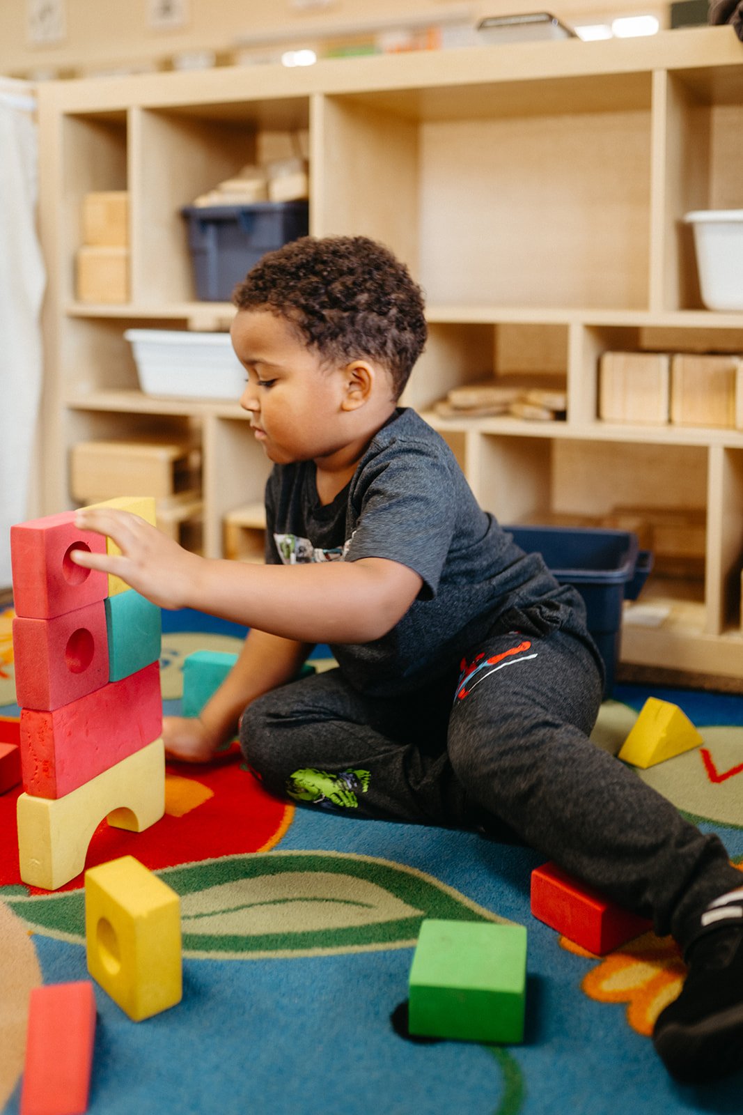 A young boy playing with colorful foam blocks on a rug in a classroom or play area.