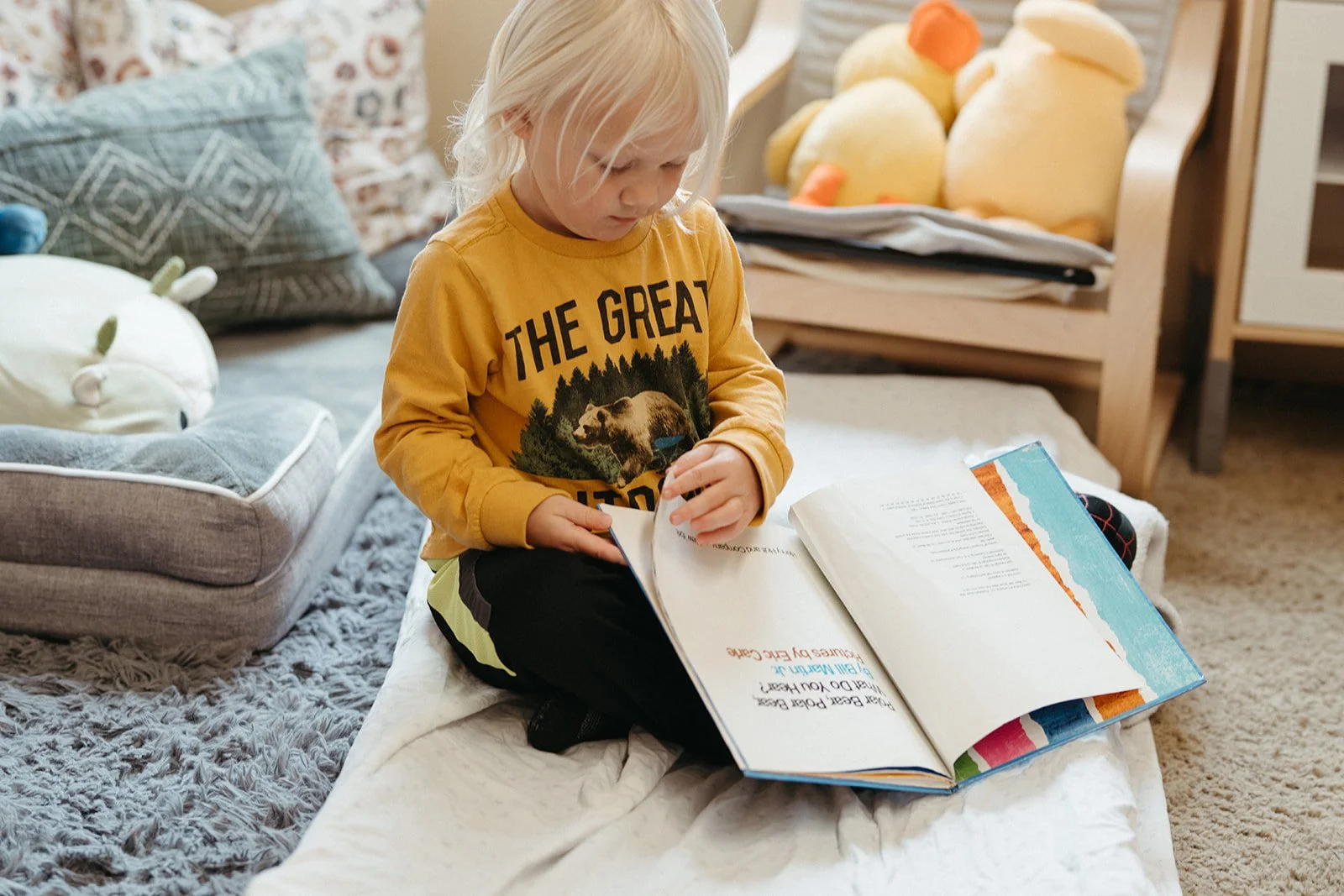 Young boy sitting on a bed, reading a large picture book. The bed has a gray fluffy blanket and stuffed pillows, with decorated pillows in the background. A wooden couch with yellow stuffed bears is also visible.