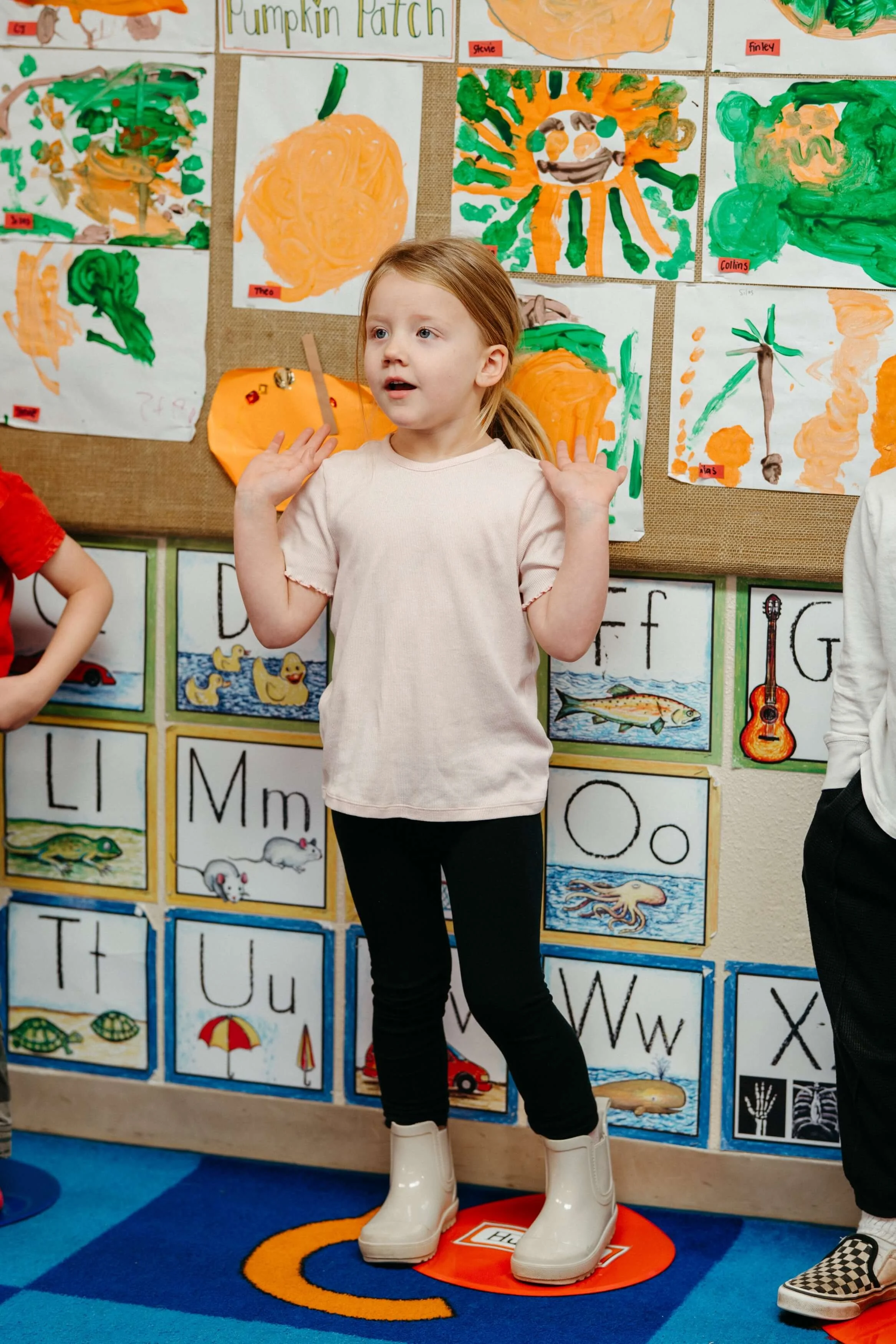 A young girl standing on an orange marker with white boots, hands raised, in front of a bulletin board displaying colorful children's artwork, including pumpkin and animal illustrations.