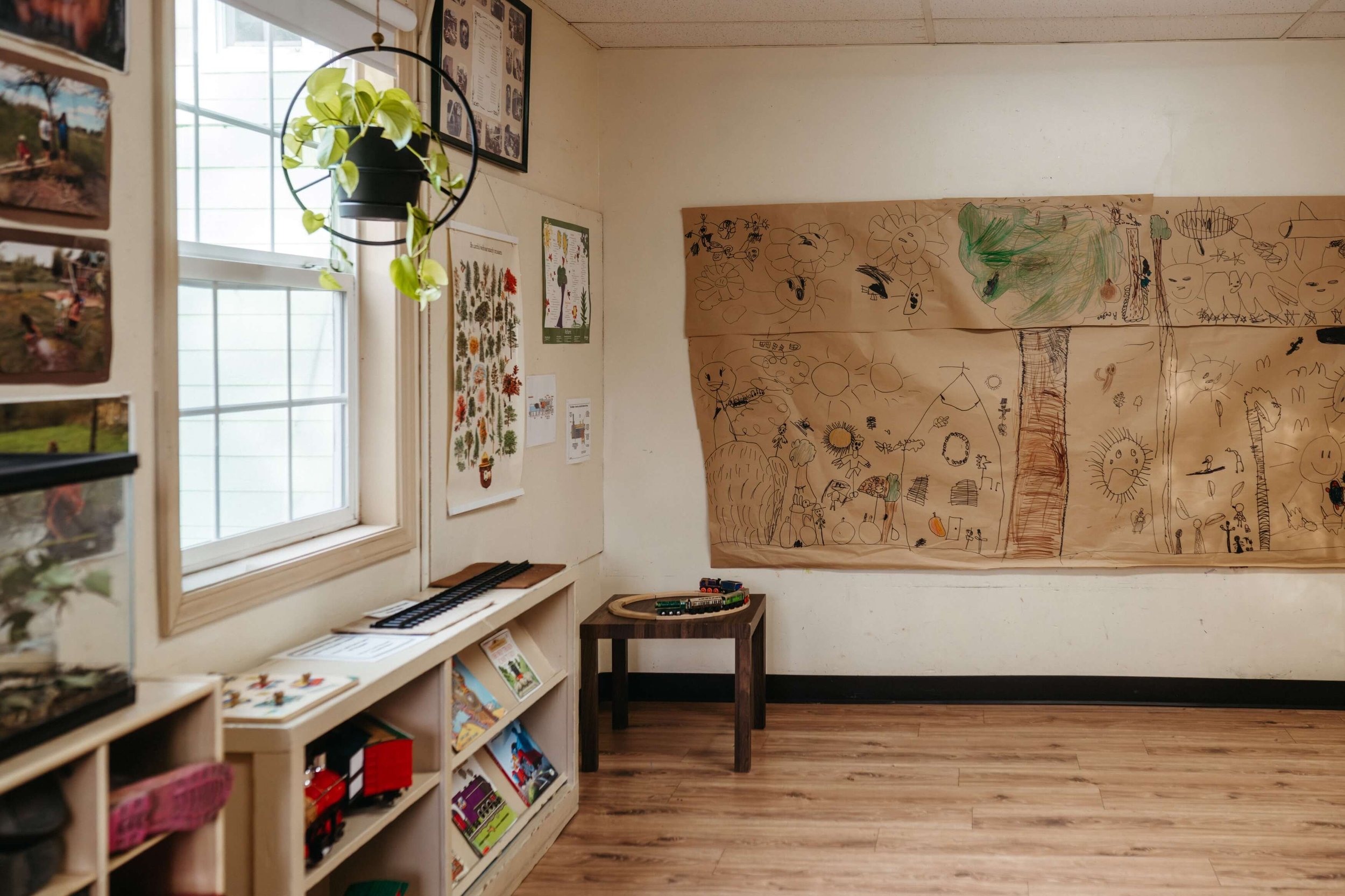 A classroom with wooden floors, a large window allowing natural light, artwork of flowers and other objects on the paper taped to the wall, and educational posters and books on shelves.