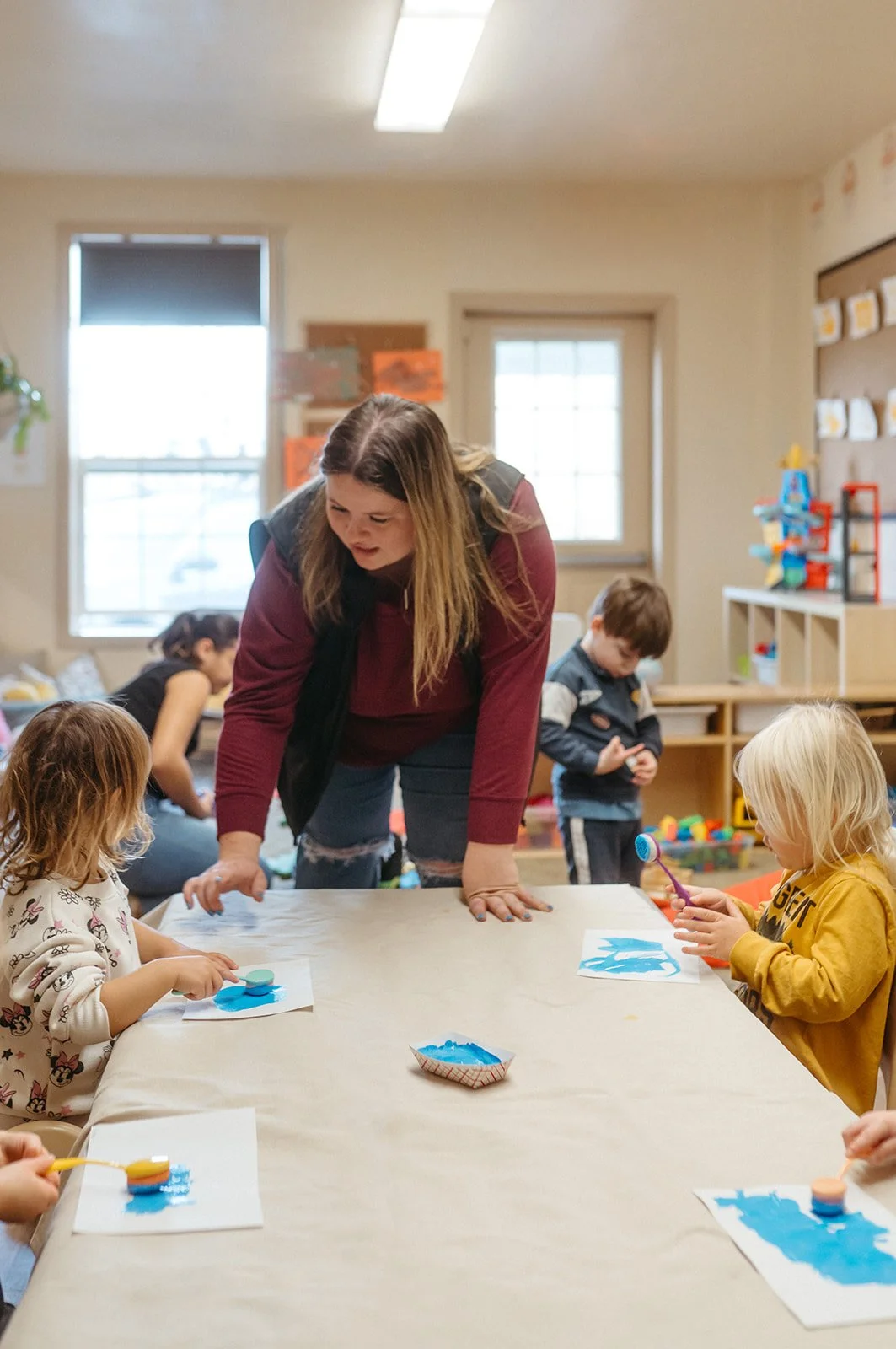 A woman is leaning over a table with young children engaged in painting activity in a classroom.