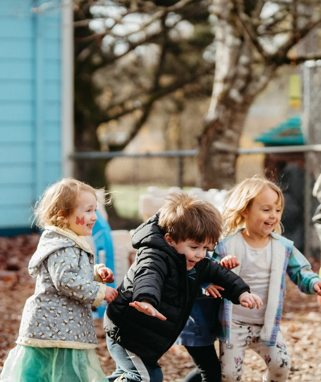 Four children playing and laughing outdoors in fall, with fallen leaves on the ground and trees in the background.