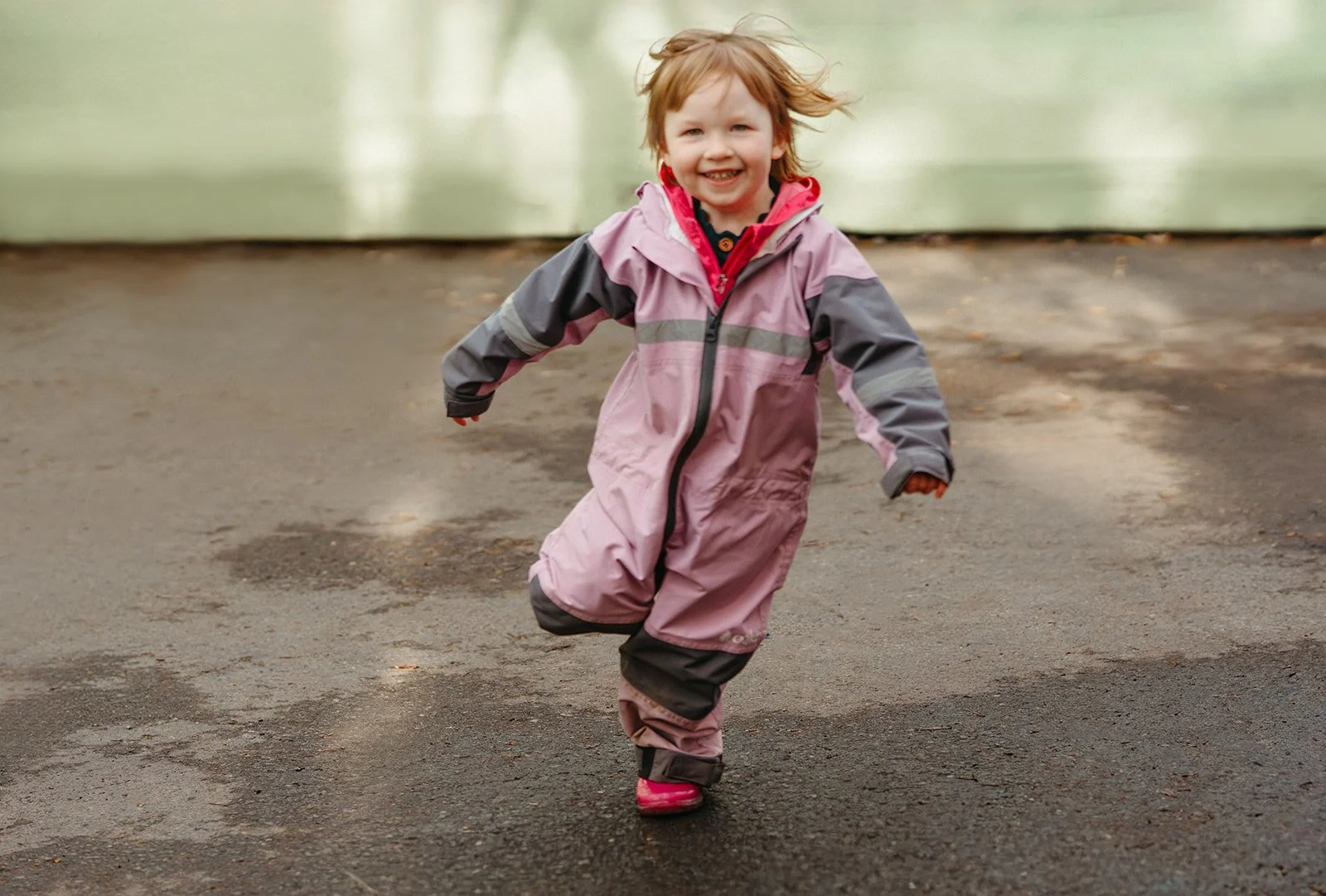Young girl running outdoors with a big smile, wearing a pink and gray winter snowsuit and pink boots.