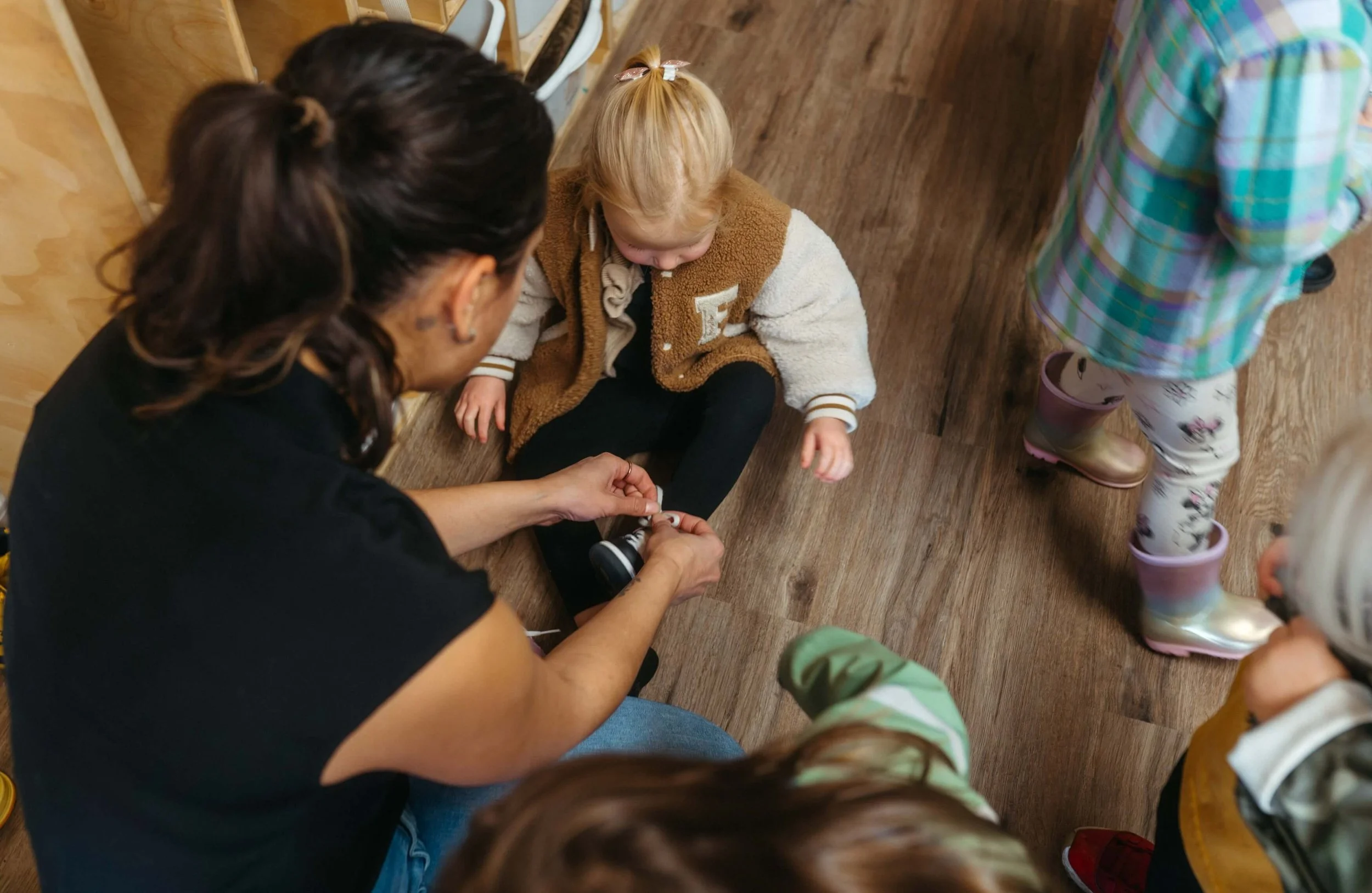 A woman is helping a young girl put on black boots, with other children standing nearby, in a room with wooden flooring and chairs.