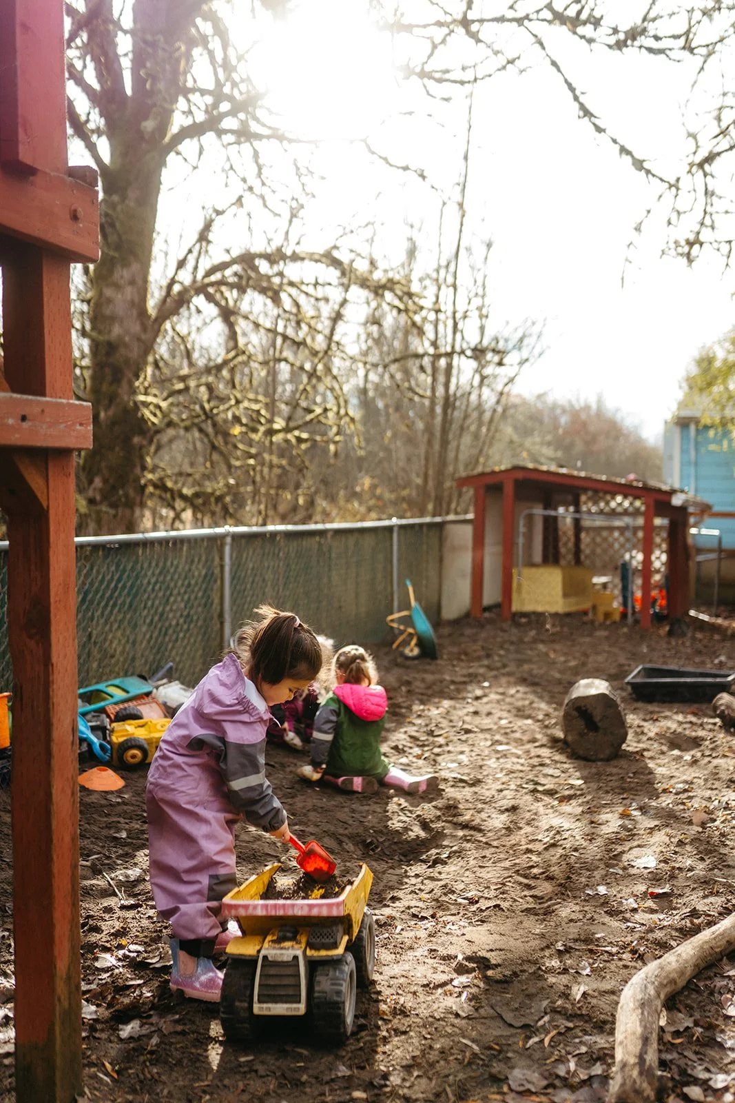Two young girls playing in a muddy outdoor play area, with toys scattered around and a small wooden shed in the background, captured in late afternoon sunlight.