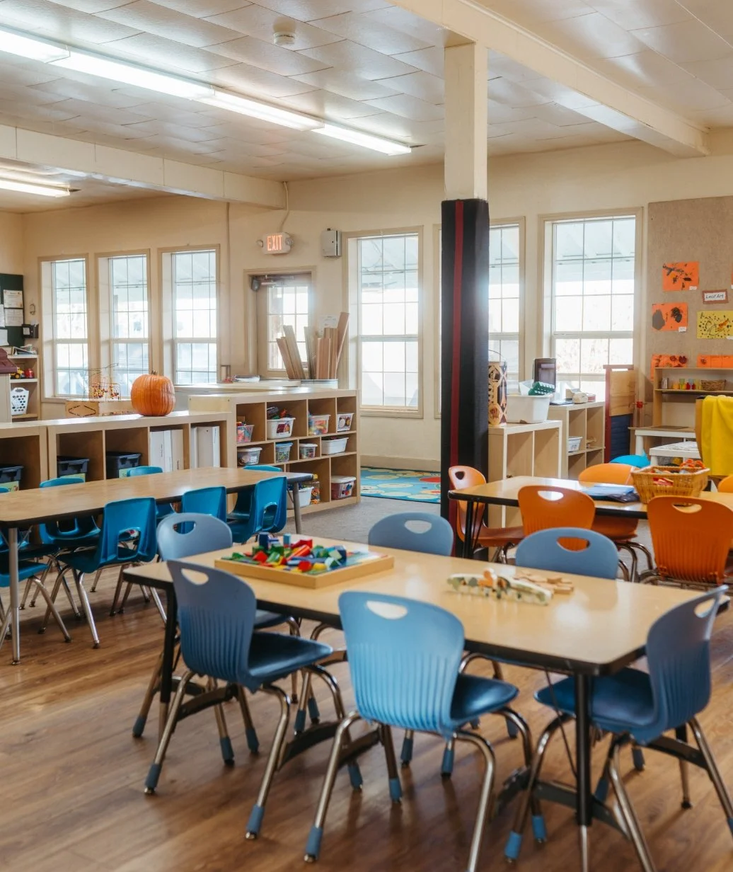 A classroom with tables, colorful chairs, educational materials, and windows letting in natural light.