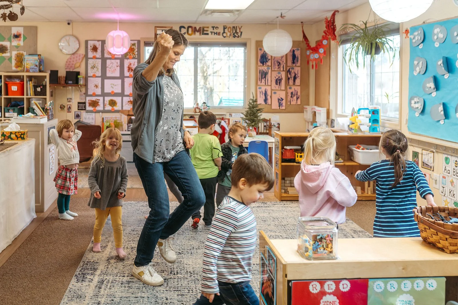 A classroom with young children and a teacher, engaging in a dance or movement activity. The room is decorated with artwork, holiday decorations, and educational materials.