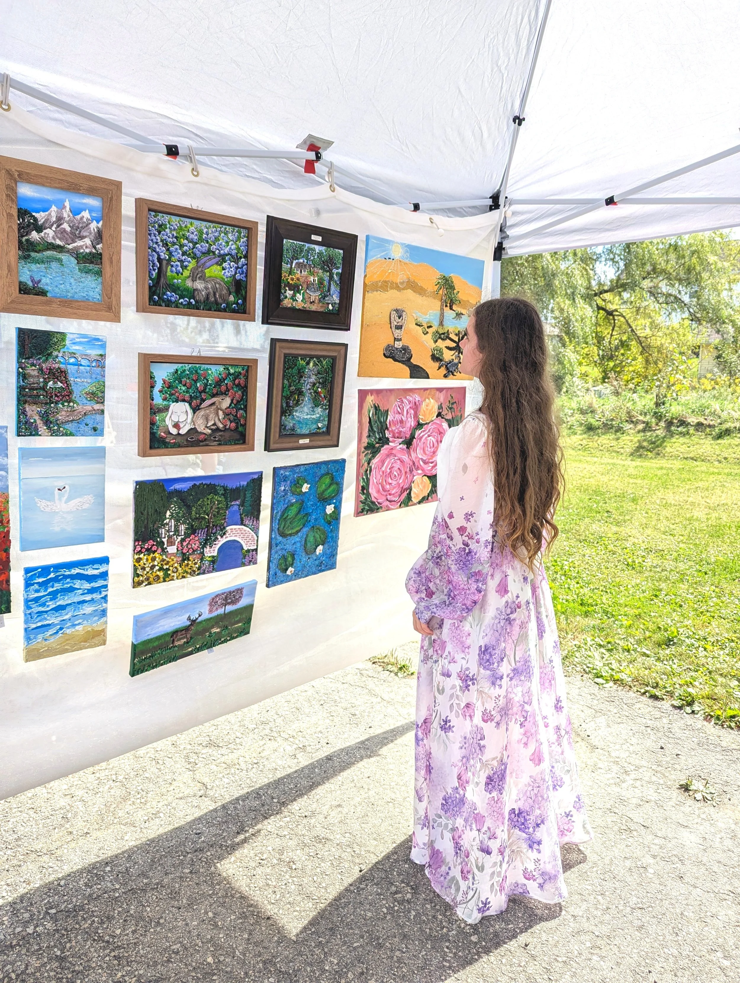 A woman in a long floral dress looking at paintings displayed on a white tent at an outdoor art fair.