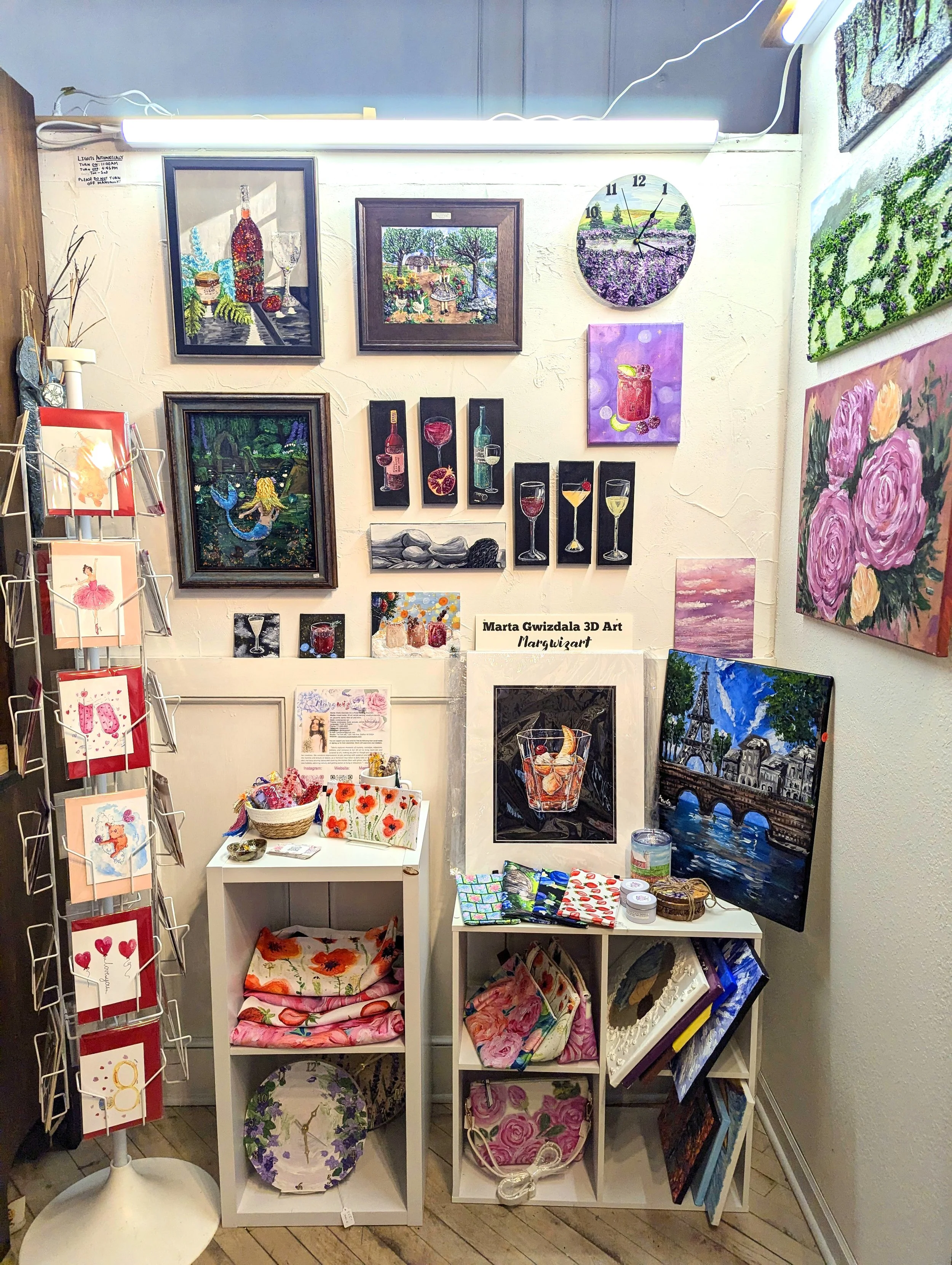 Art gallery display with paintings, wall clock, greeting cards on a stand, and small decorative items on shelves.