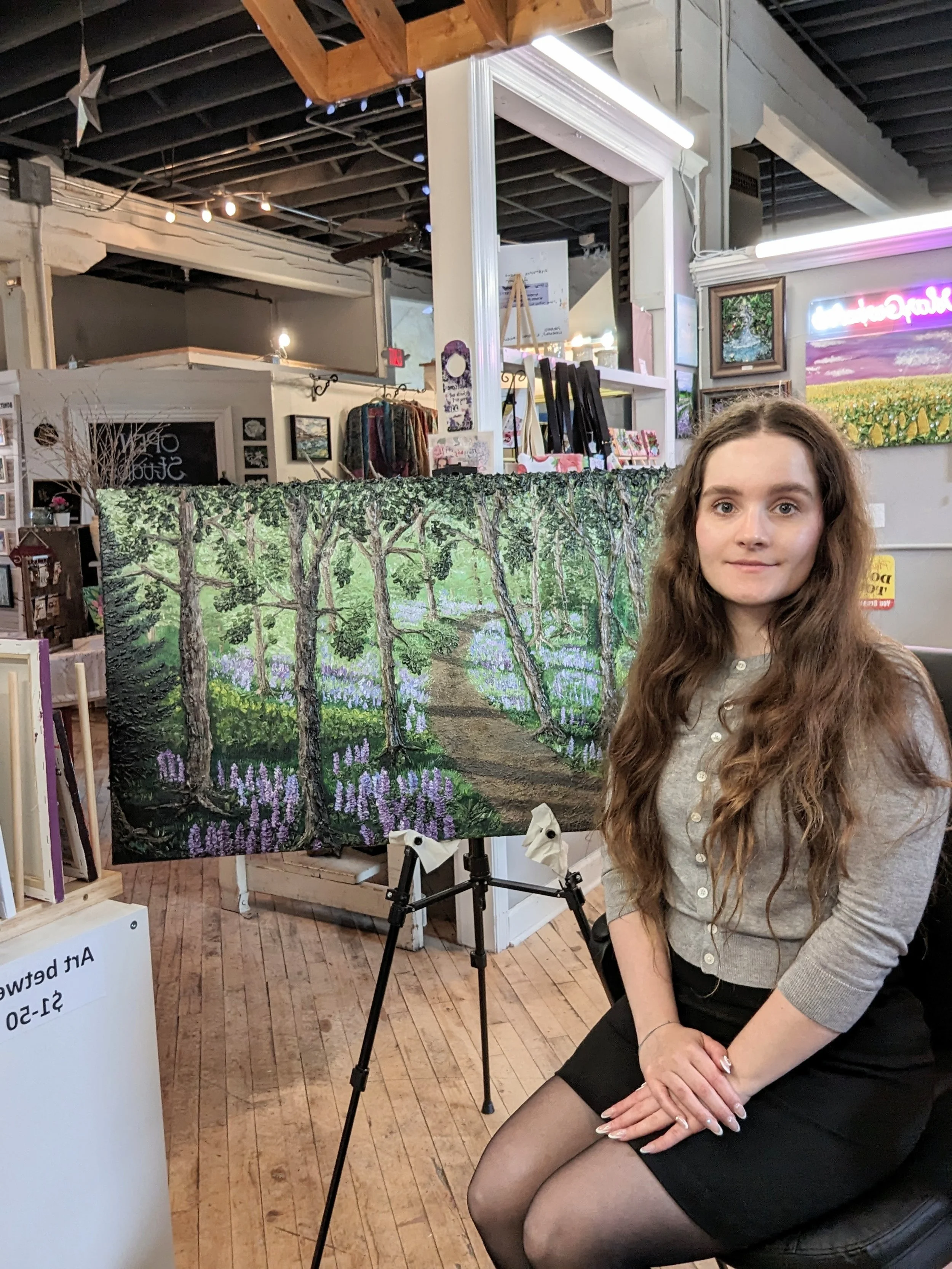 A young woman with long, wavy brown hair and light skin sitting in an art gallery or store, next to a framed landscape painting of a tree-lined path and a field of purple flowers.
