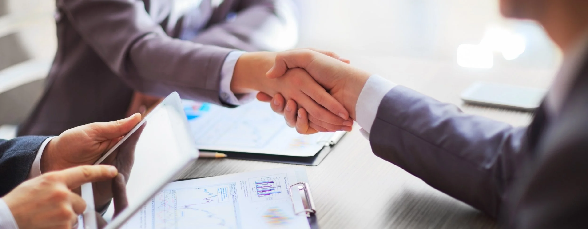 Close-up of two people shaking hands in a business setting, with documents, charts, and a tablet on the table.