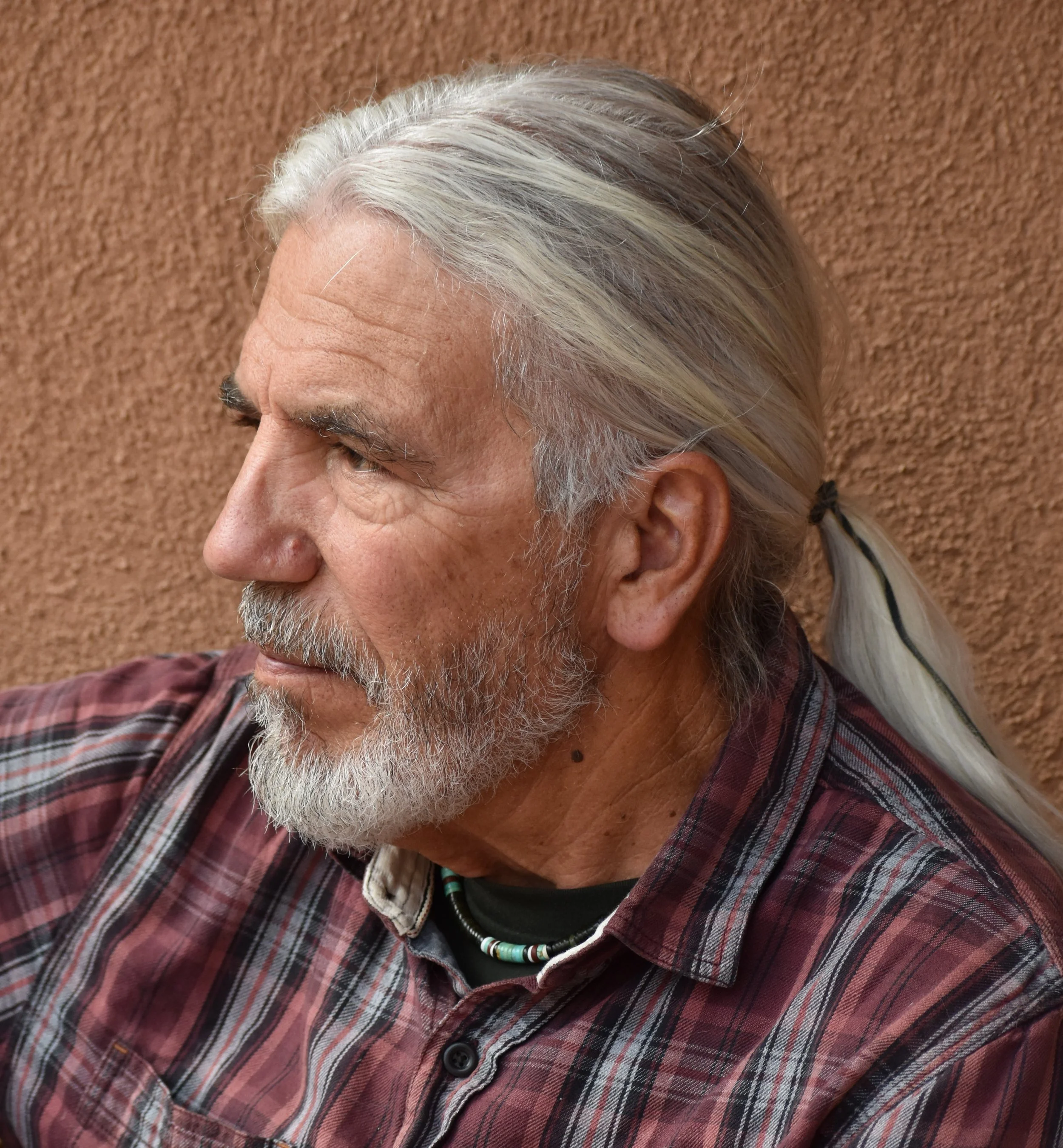 Profile of an older man with long gray hair tied in a ponytail, a gray beard, wearing a plaid shirt and beaded necklace, against a textured orange wall.