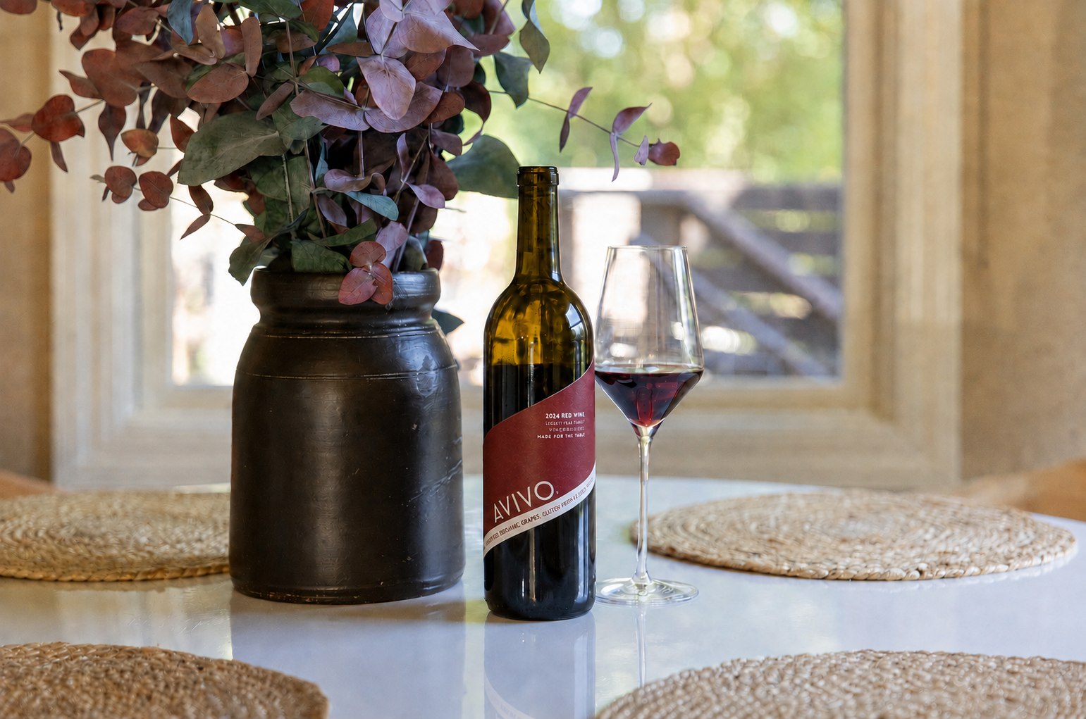 A table with a vase of purple and green leaves, a bottle of red wine, and a wine glass filled with red wine in a room with natural light and a window in the background.