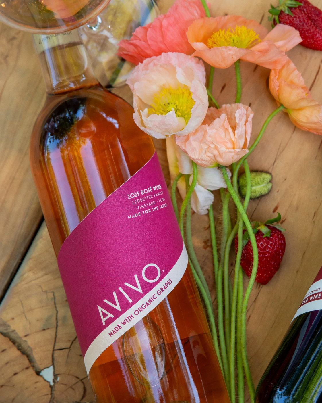 A bottle of rosé wine labeled 'AVIVO' next to pink and peach poppy flowers and strawberries on a wooden surface.