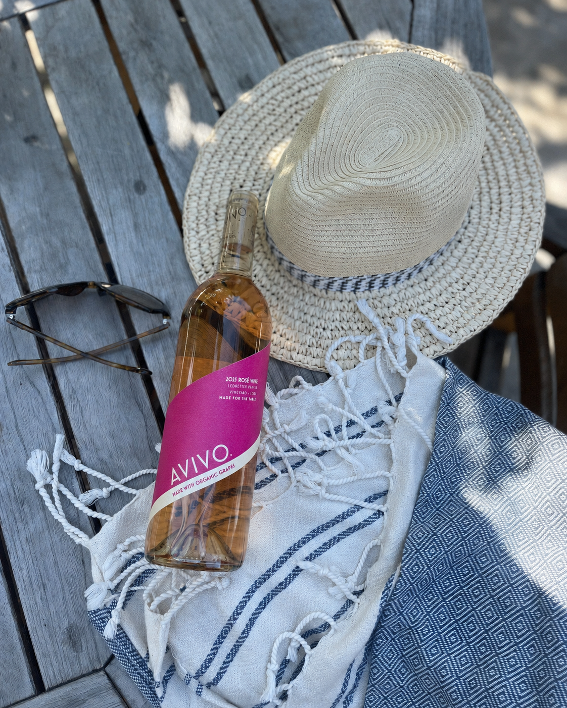 A straw sun hat, a bottle of rosé wine labeled 'AVIVO,' sunglasses, and a patterned fabric on a weathered wooden surface, with sunlight and shadows.