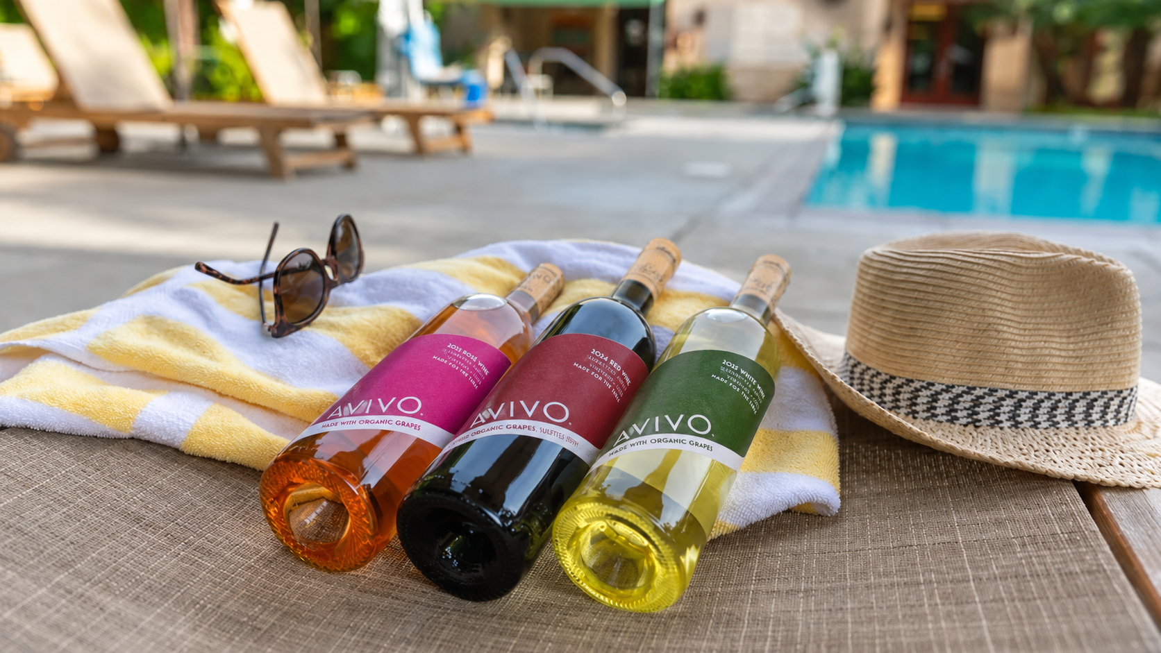 Three bottles of colorful beverages, a pair of sunglasses, a straw sun hat, a yellow and white striped towel, and a swimming pool in the background.