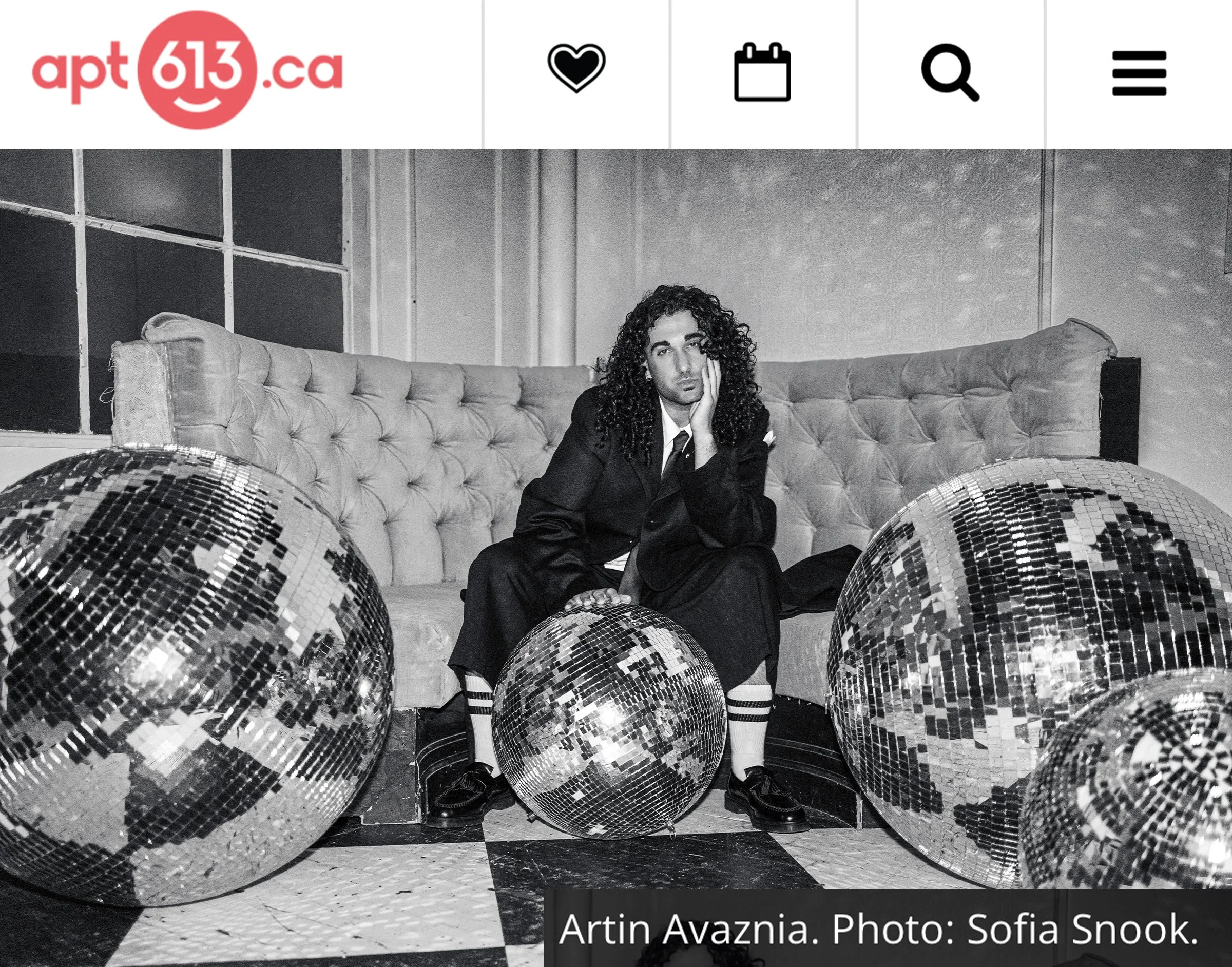 Black and white photo of a man with curly hair sitting on a vintage tufted sofa, surrounded by disco balls, with a background of wall panels.