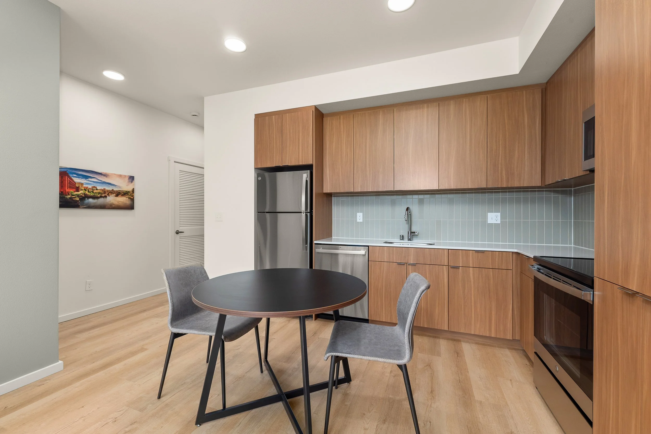 Modern kitchen with wooden cabinets, stainless steel refrigerator, microwave, and oven. Small round black dining table with two gray upholstered chairs. Light wood flooring and a picture on the white wall.