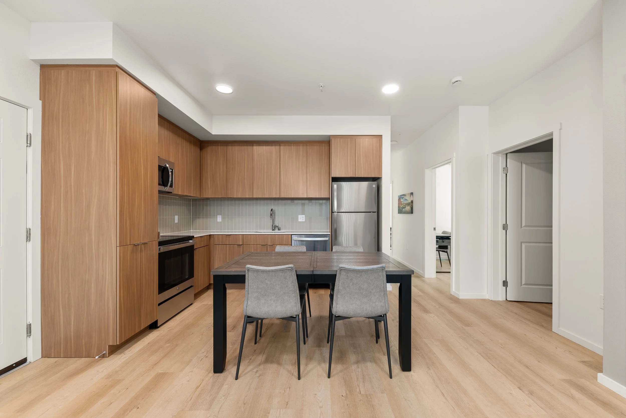 Modern kitchen with wooden cabinets, stainless steel appliances, a rectangular dining table with four gray chairs, and a light wood floor.