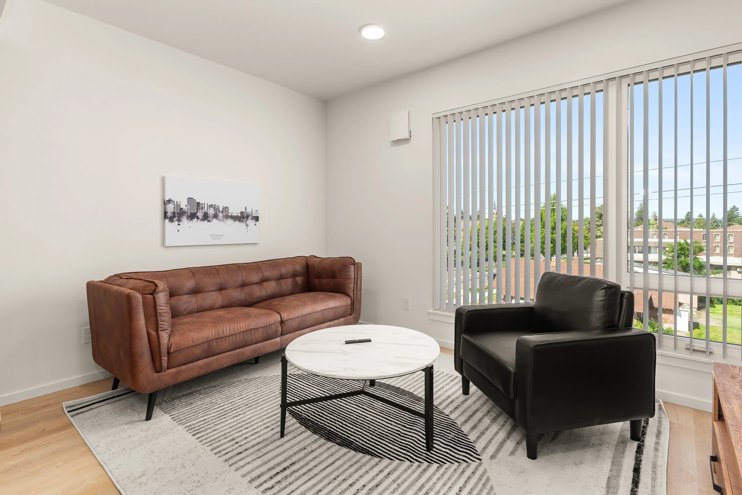 Living room with brown leather sofa, black armchair, white marble coffee table, striped rug, window with vertical blinds, and cityscape view outside.