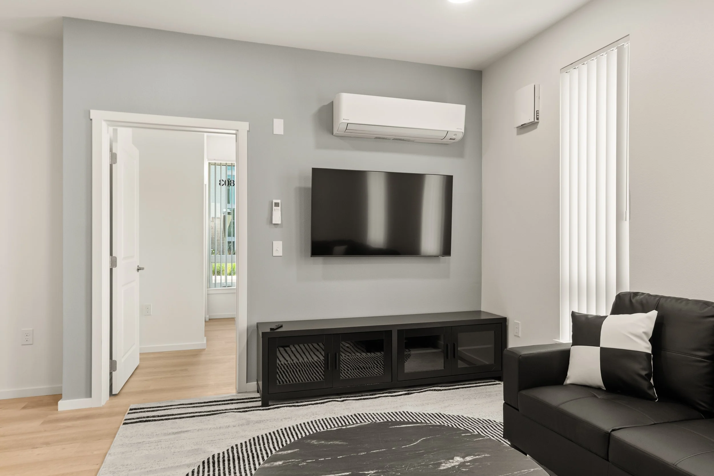 Living room with gray walls, black TV on wall, black TV stand, air conditioning unit above TV, window with vertical blinds, black leather sofa with black and white pillows, striped rug, and wooden flooring.