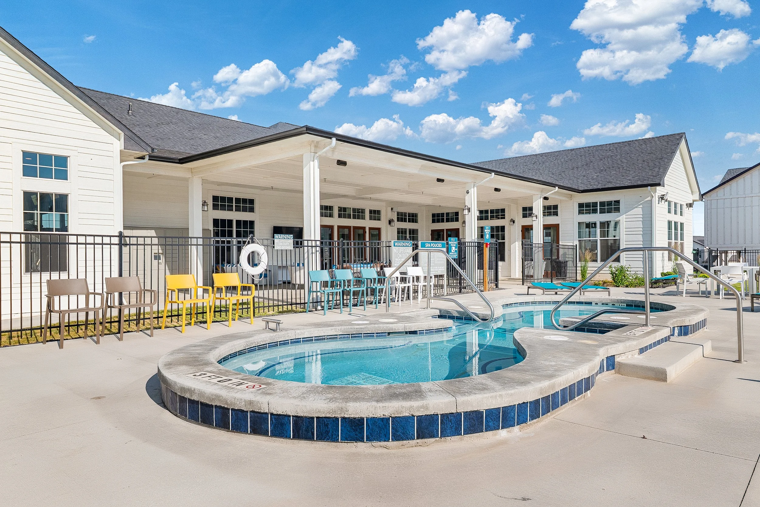 Swimming pool and hot tub area at a residential complex with white buildings, blue sky, and clouds in the background.