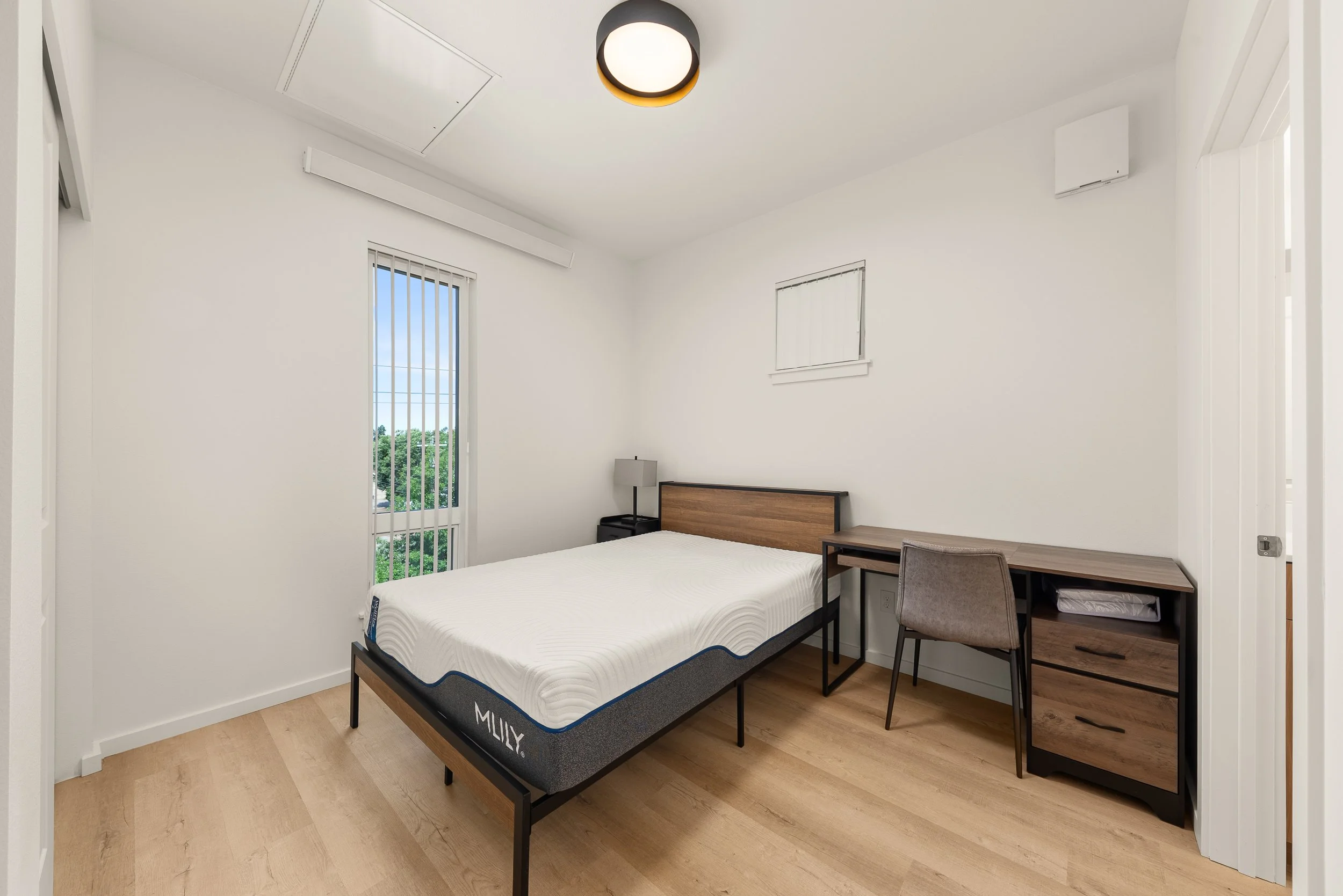 A minimalist bedroom with white walls and a wooden floor, a bed with a white mattress, a small dark nightstand with a lamp, a wooden desk with a gray chair, and a window with vertical blinds.