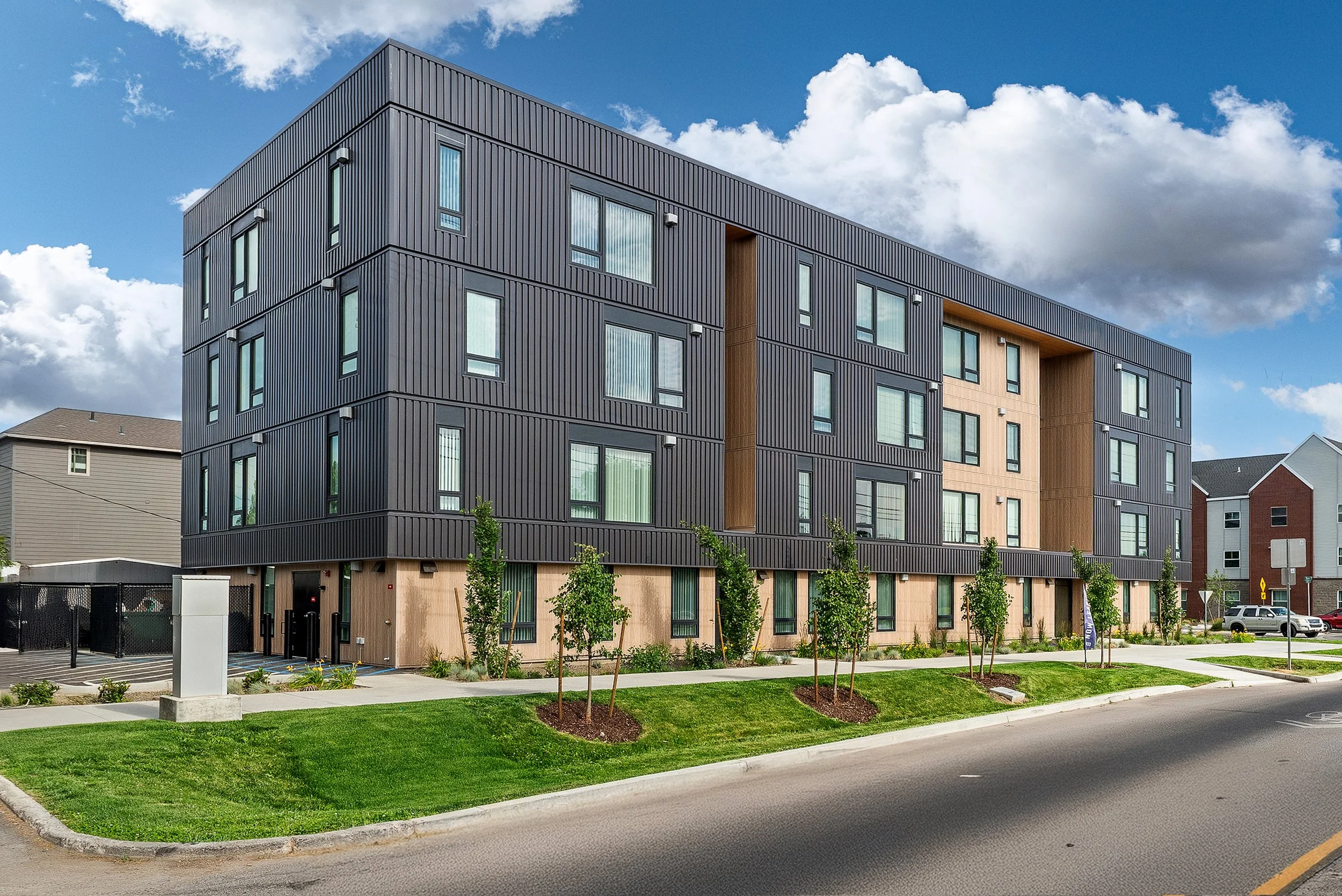 Modern multi-story apartment building with black and beige exterior, trees, sidewalk, and parked cars in a suburban area under a partly cloudy sky.