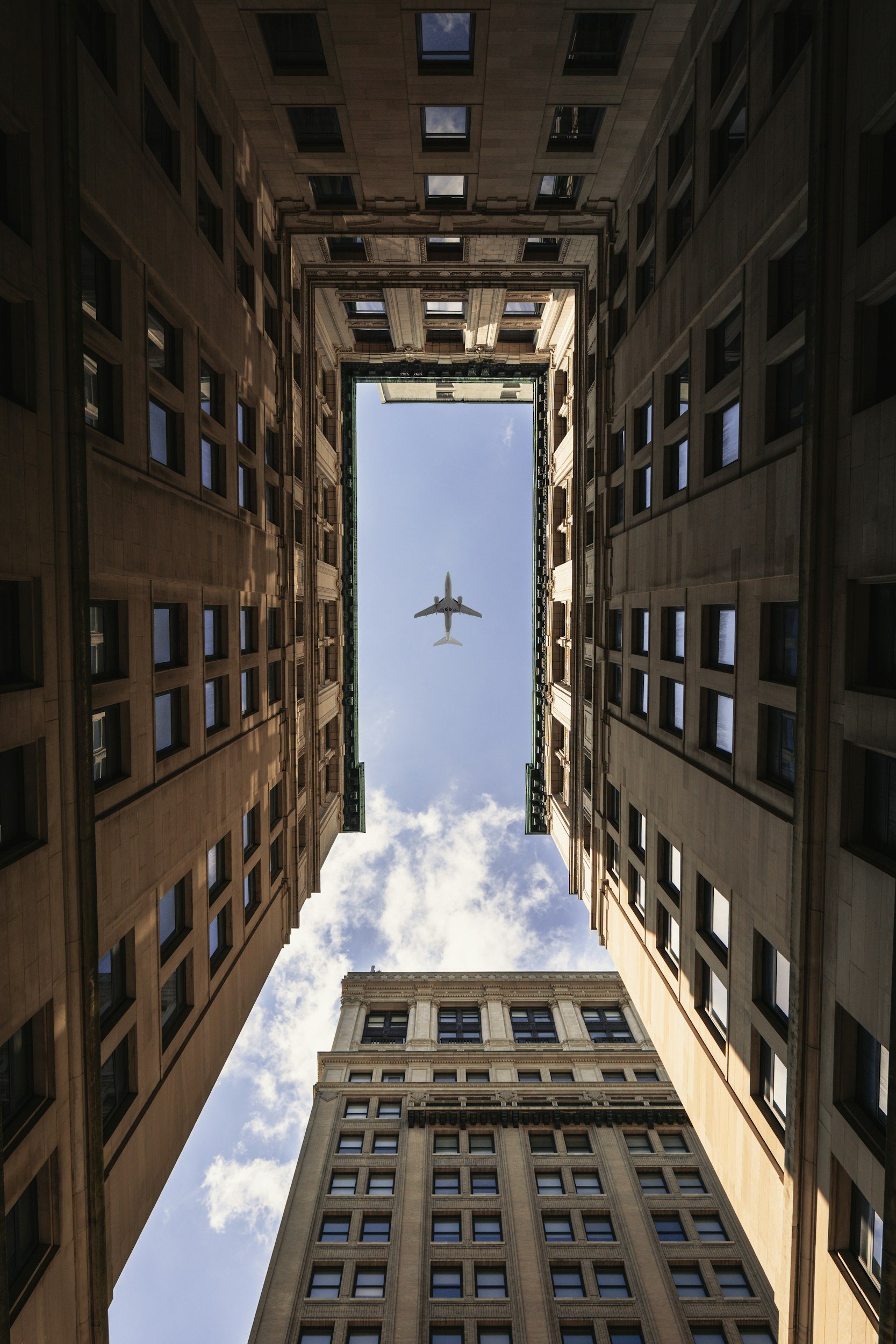 An upward view of tall buildings framing a blue sky with an airplane flying overhead.