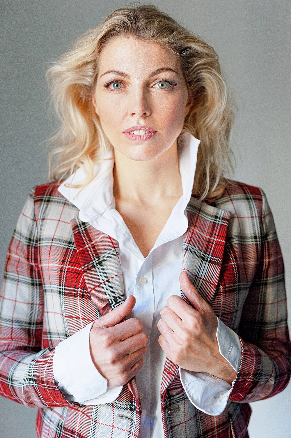 A woman with blonde hair and blue eyes looking at the camera, wearing a plaid blazer over a white shirt, holding the lapels of her blazer.