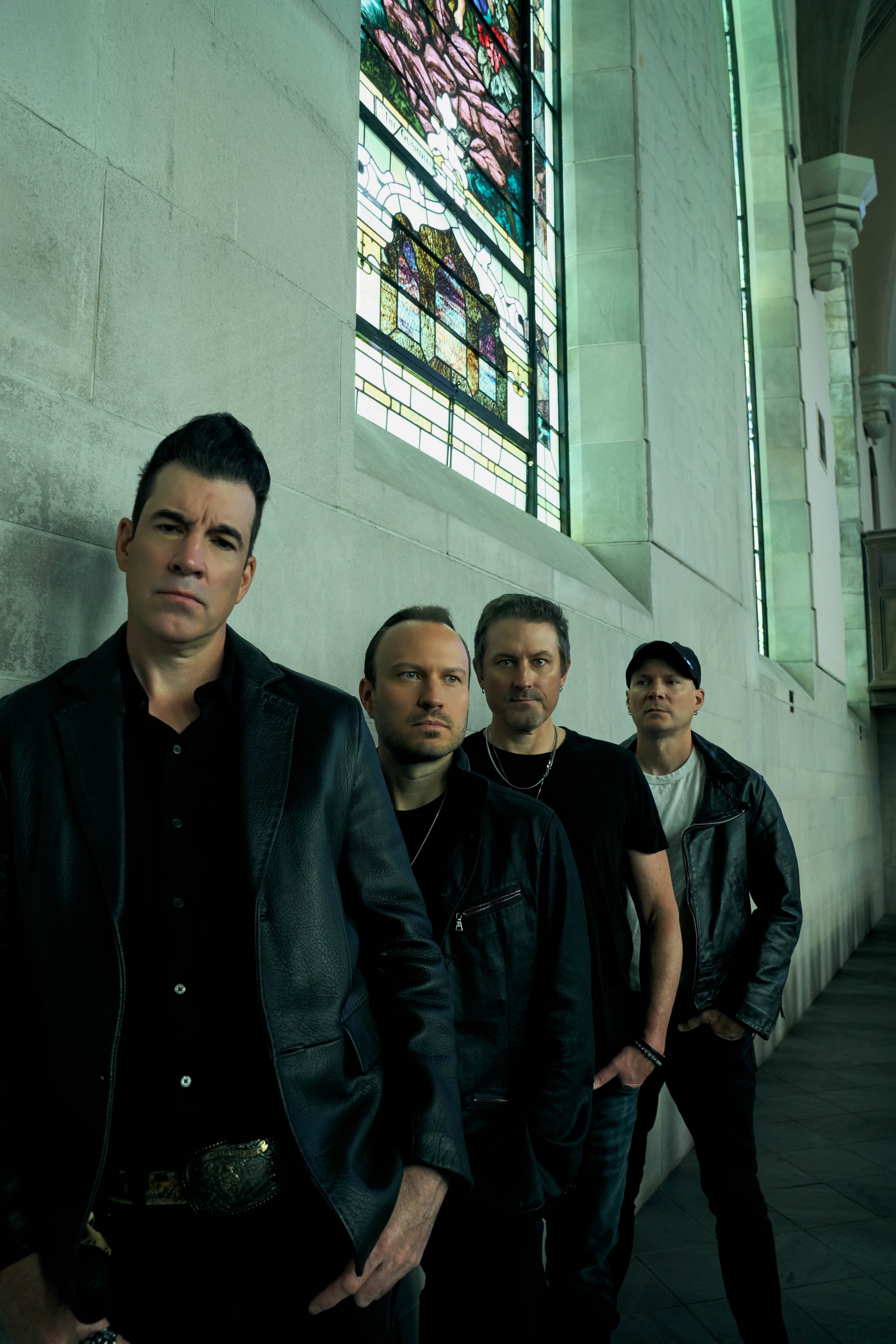 A group of four men standing in a line inside a building with stained glass windows and stone walls. The four men are the band members of Theory of a Deadman - Tyler Connolly, Dean Back, Dave Brenner, and Joe Dandeneau.