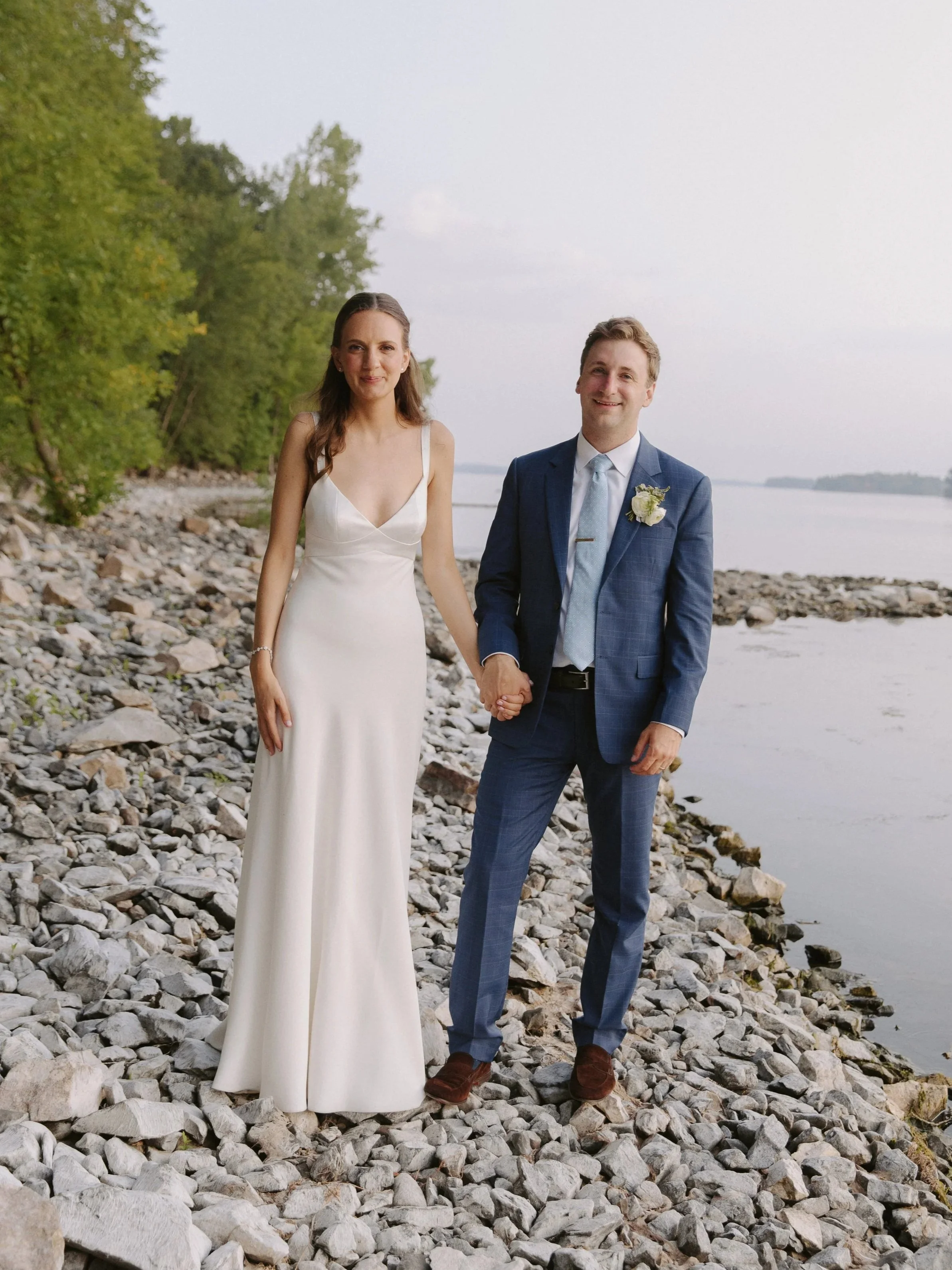 A couple in wedding attire standing on a rocky shoreline by a lake, holding hands and smiling. The bride wears a white gown, and the groom wears a blue suit with a boutonniere.
