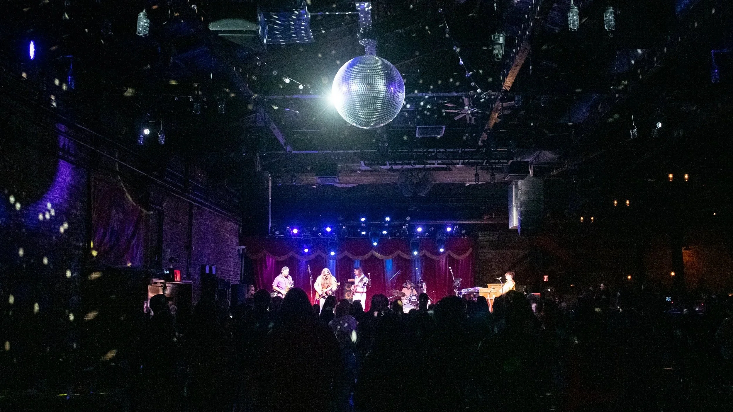 A live music performance on stage with a guitarist, bassist, drummer, and keyboardist, under purple and blue stage lights, in a dimly lit venue with an audience watching, and a disco ball hanging from the ceiling.