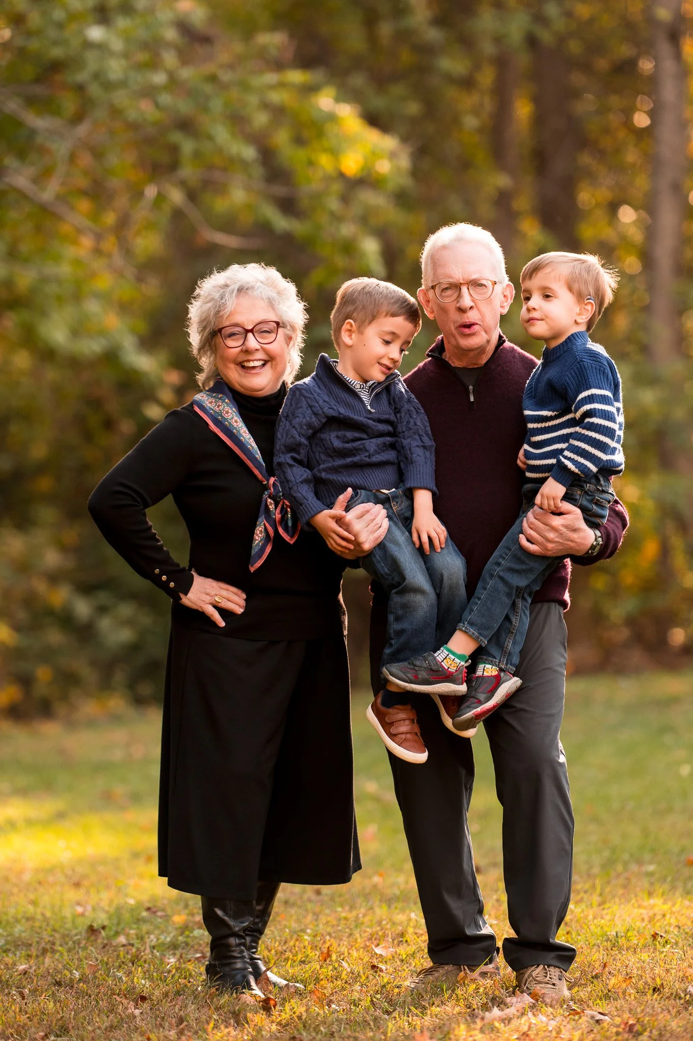 A happy, multi-generational family of four outdoors in a park during fall, with trees and fallen leaves in the background. The grandparents are holding their two young grandchildren, all smiling and enjoying the moment.