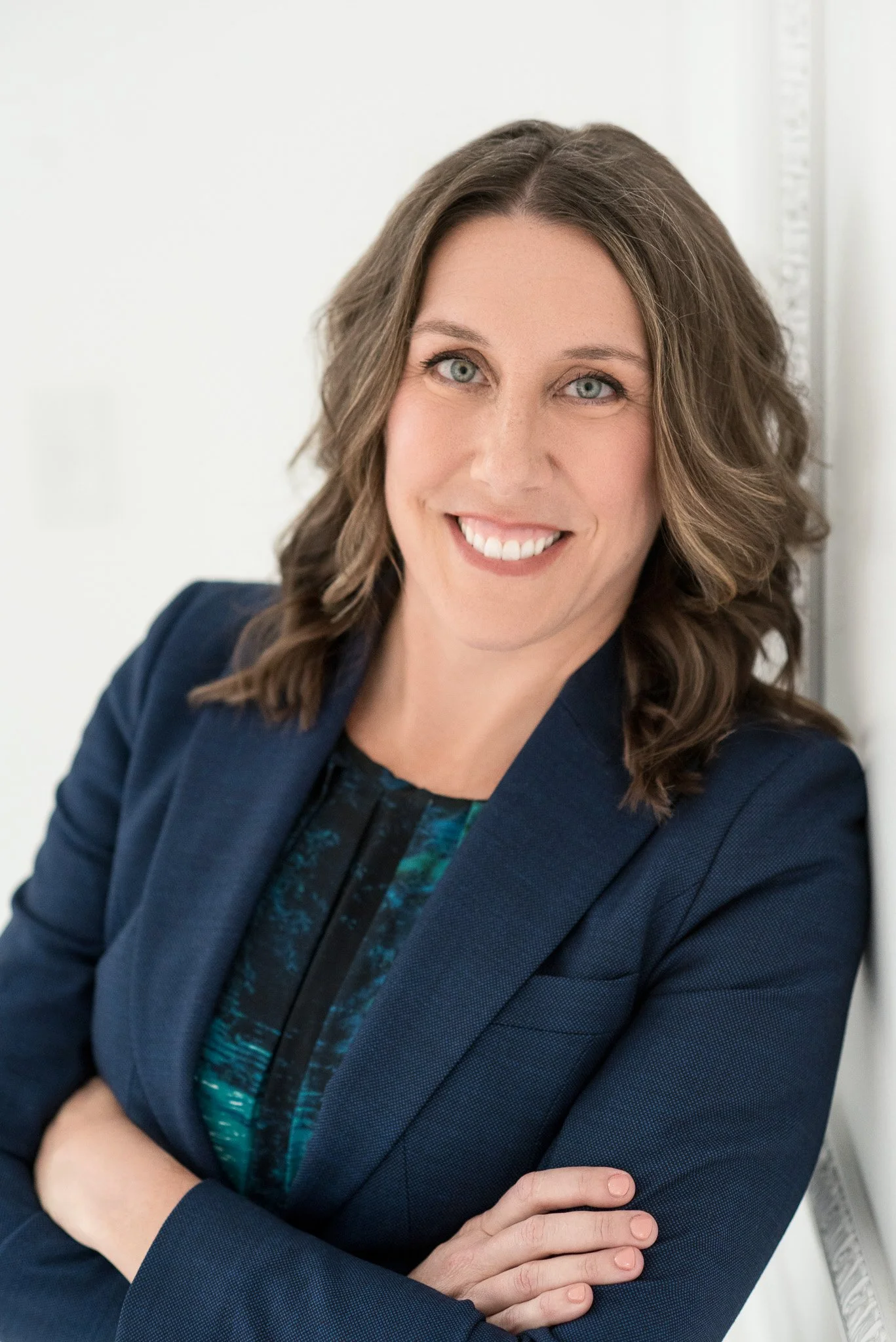 A woman with shoulder-length wavy brown hair, blue eyes, wearing a navy blazer and patterned blouse, smiling and crossing her arms against a white background.