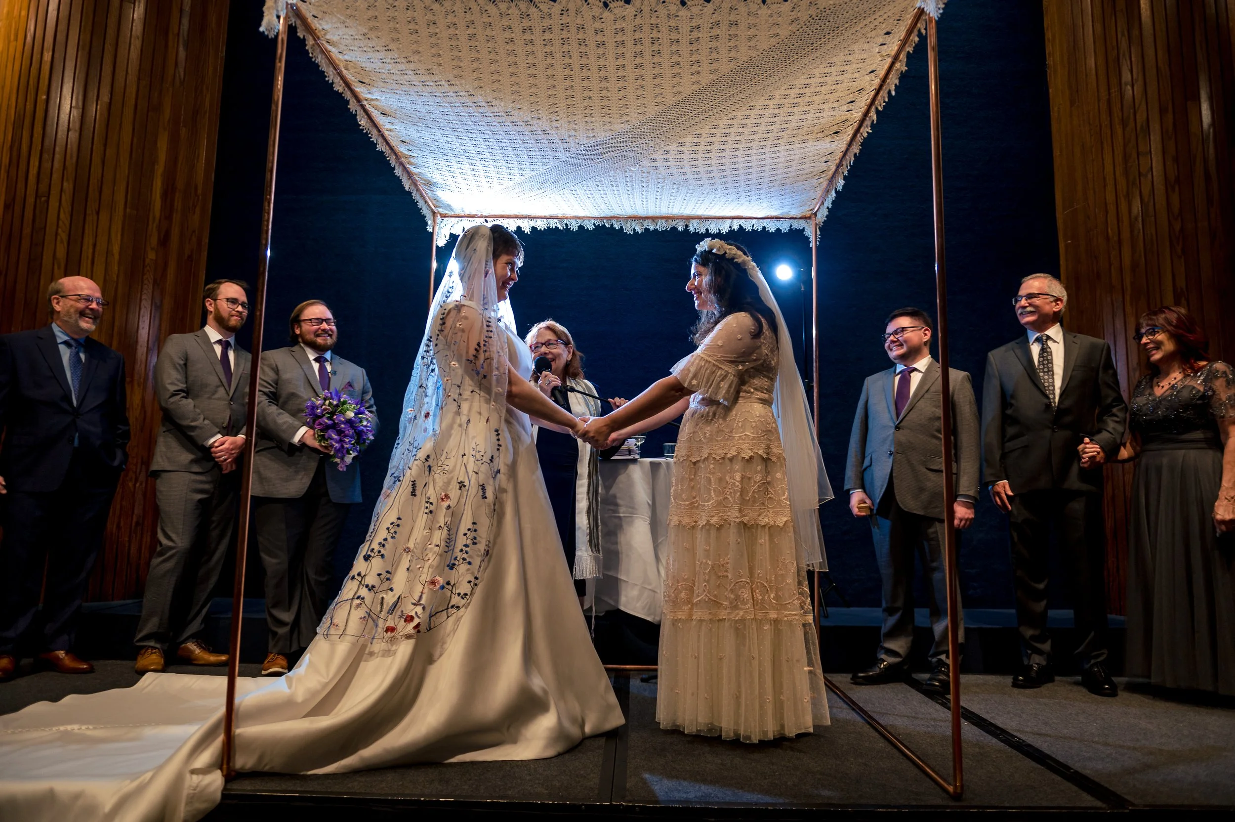 A same-sex wedding ceremony taking place under a canopy, with two women holding hands and exchanging vows, surrounded by officiants, wedding party members, and guests, all smiling and dressed formally.