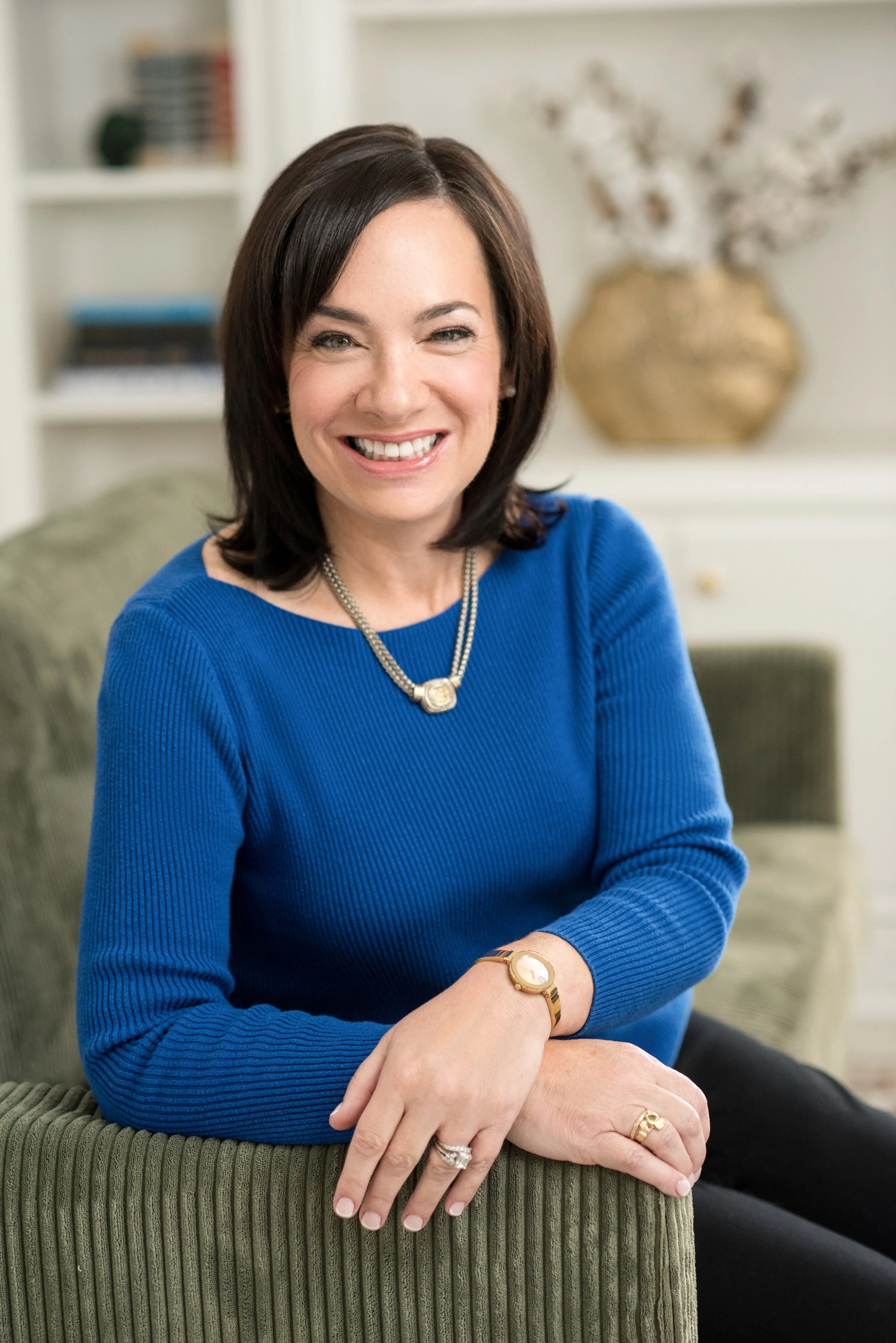 A woman with dark brown hair in a bob cut, smiling and wearing a royal blue long-sleeve top, sitting on a green upholstered armchair in a living room. She accessorizes with a gold watch, a pearl necklace, and rings on her fingers. In the background, 