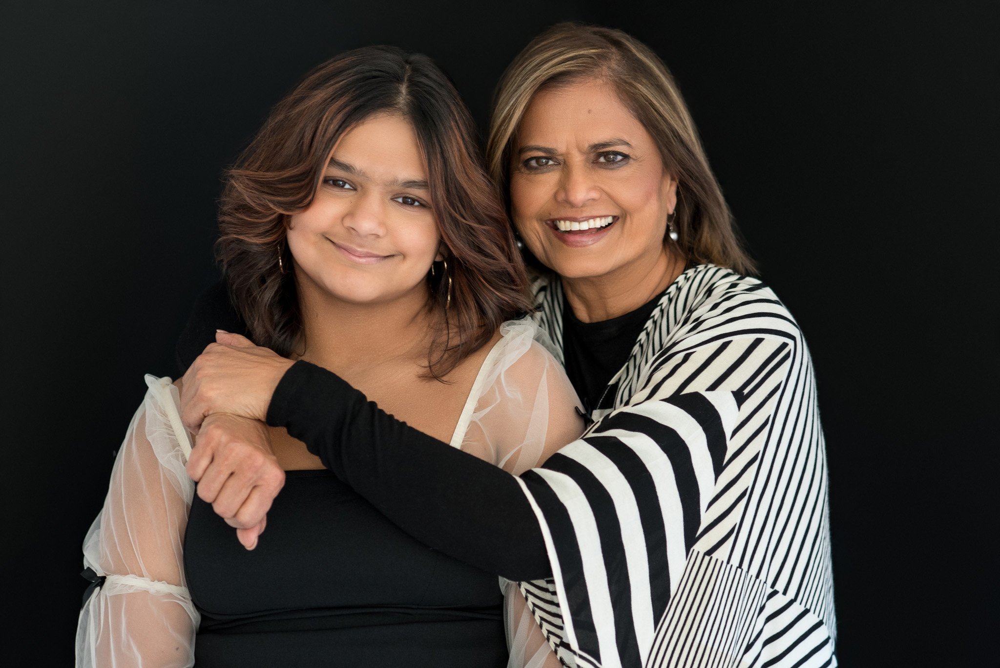 A woman and a young girl are smiling and hugging against a black background.