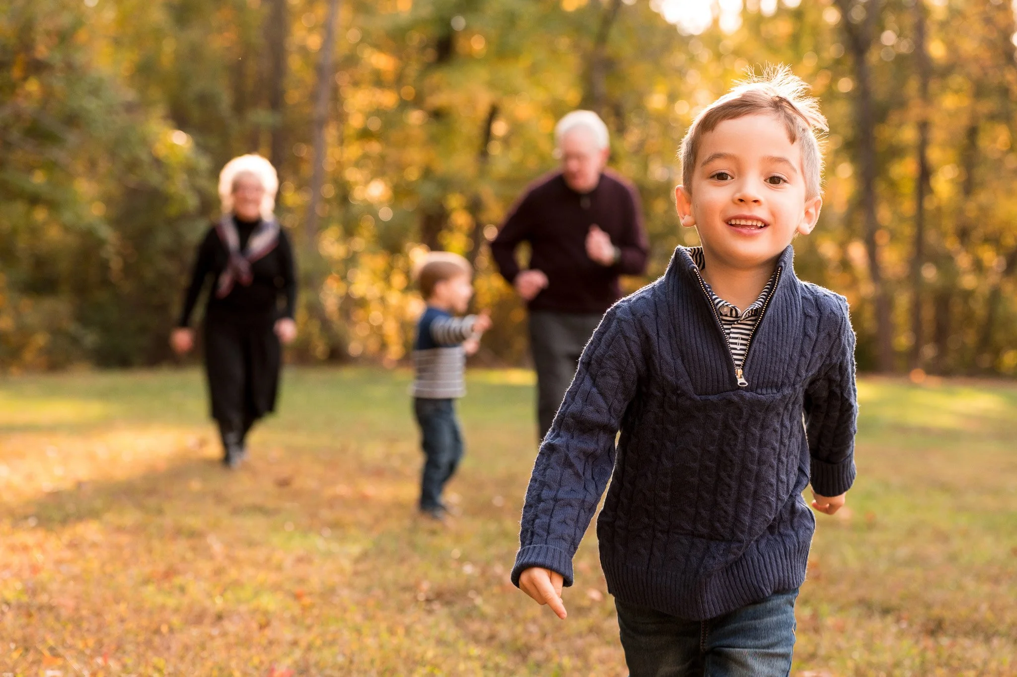 A young boy runs toward the camera with a smile, outdoors in a park during fall, with trees and other children and adults in the background.