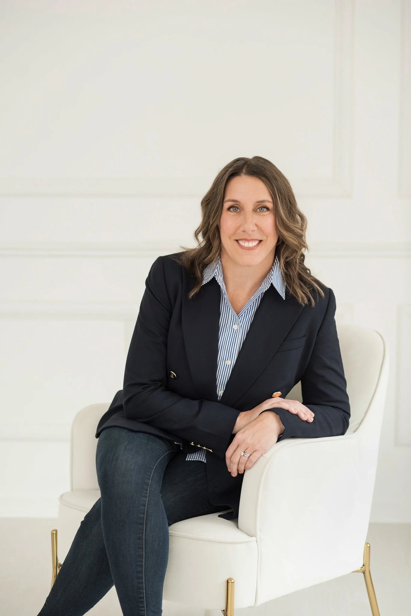 Smiling woman with wavy brown hair, wearing a dark blazer over a blue-striped shirt, sitting on a white chair in a bright room.