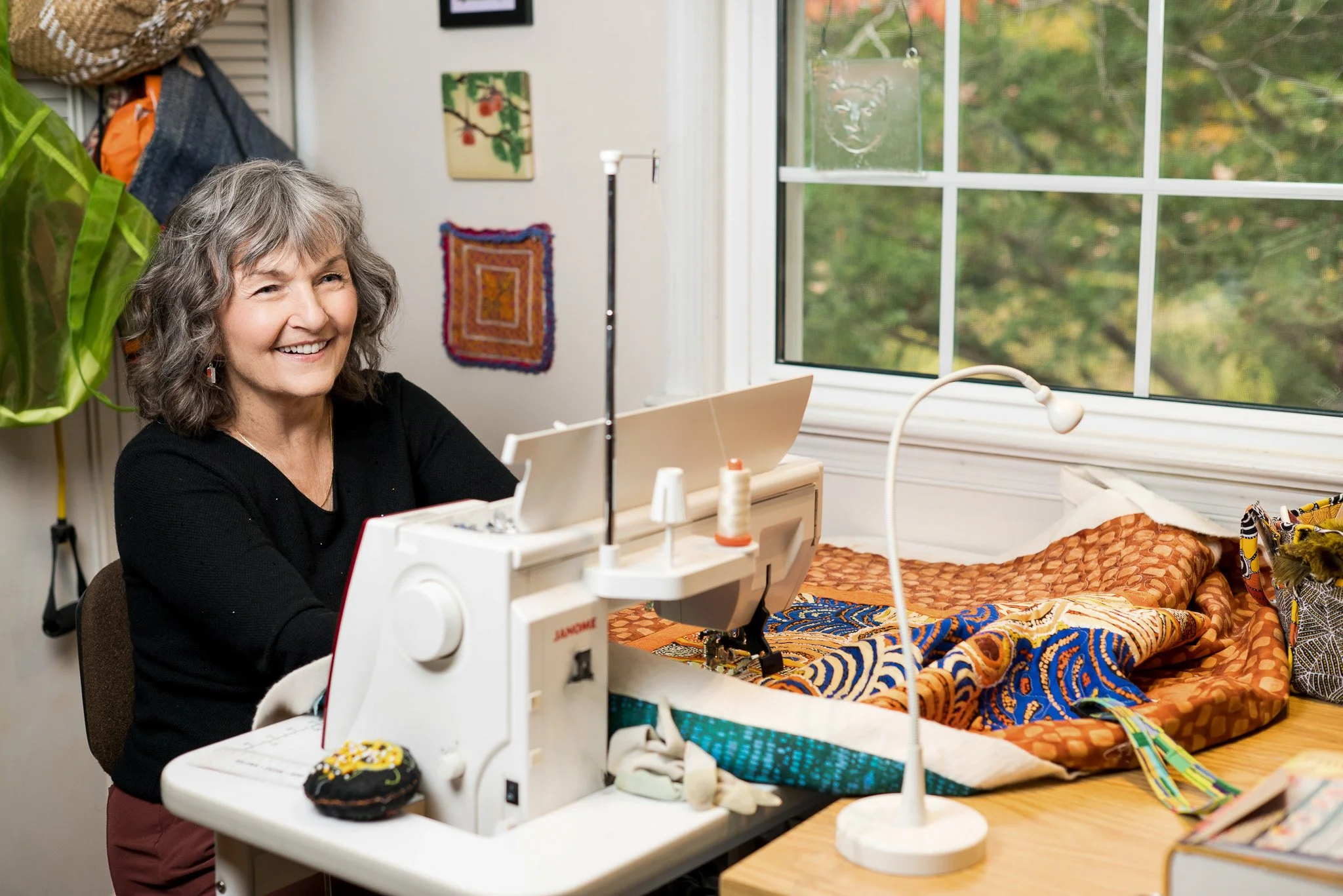 A woman with gray hair smiling while sewing fabric on a sewing machine near a window with a view of trees outside.