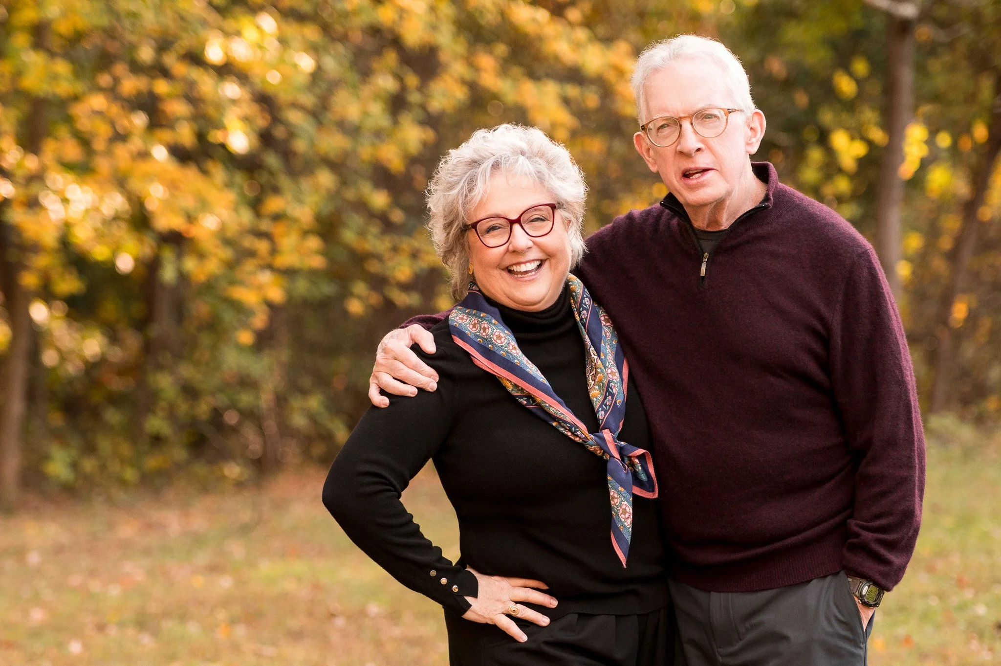 An elderly couple smiling outdoors in a park with autumn foliage, the woman has short gray hair and glasses, and is wearing a black top with a colorful scarf, the man has short white hair and glasses, and is wearing a maroon sweater.