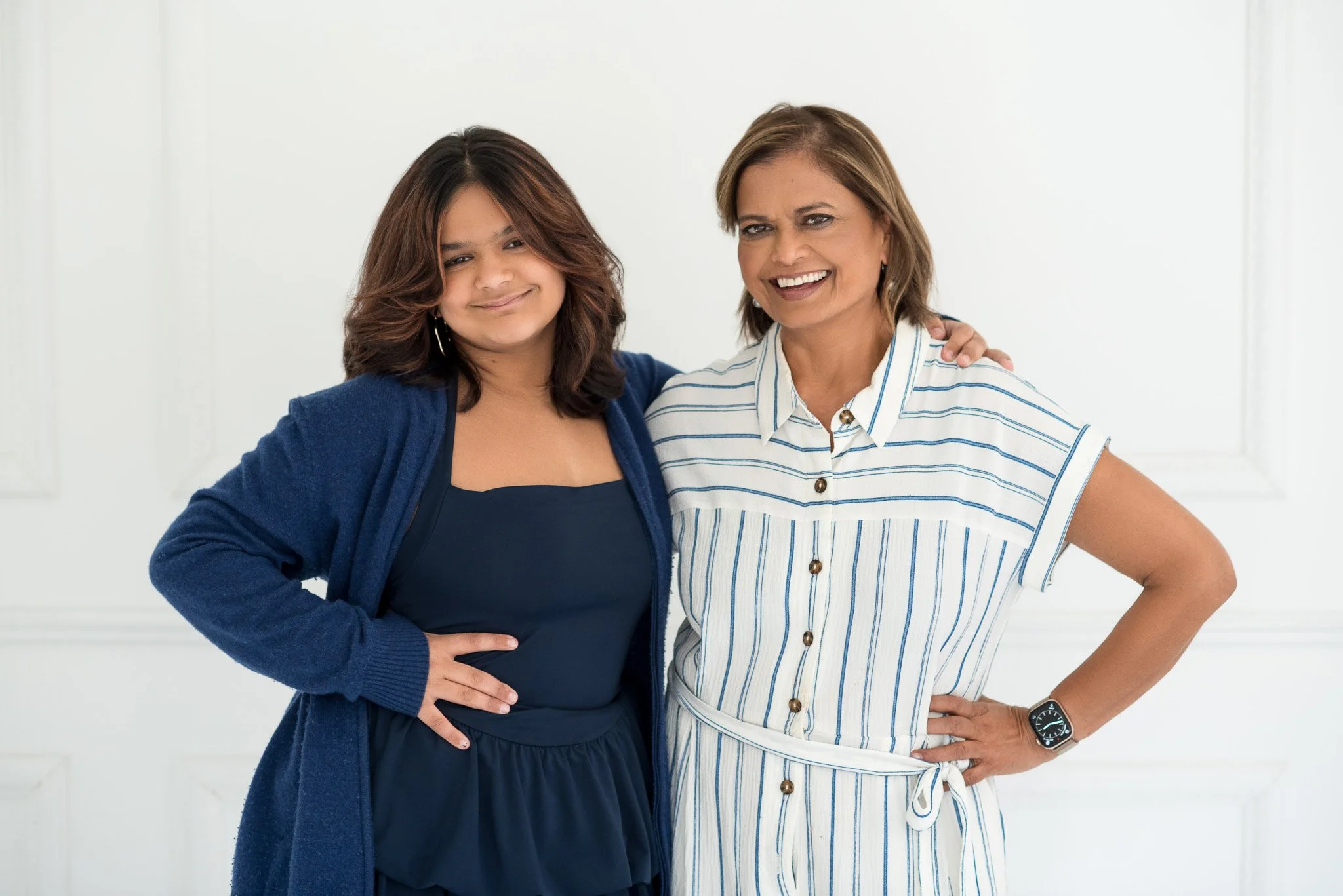 Two women standing side by side, smiling, with one arm around each other in a bright room with white walls.
