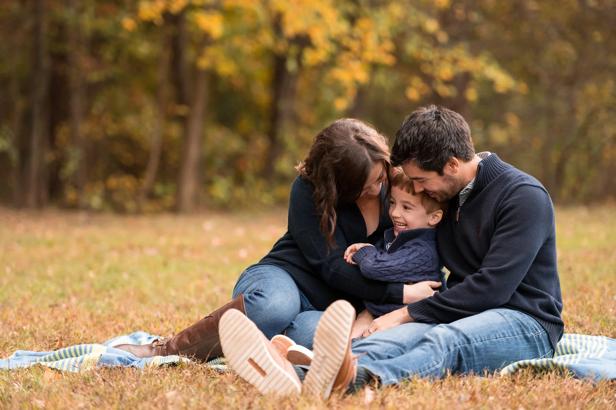 Family of three sitting on a blanket in a park with autumn foliage, smiling and playing together.