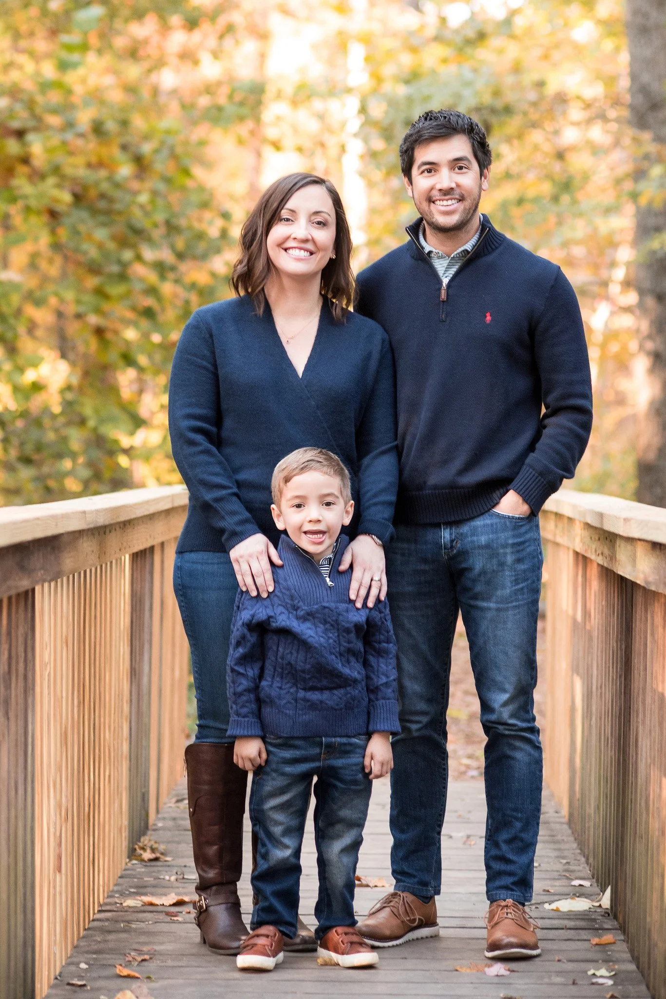 A family of four standing on a wooden bridge outdoors with autumn foliage in the background. The mother and father are standing behind their young son, who is in front of them. All are smiling and dressed in casual fall clothing.