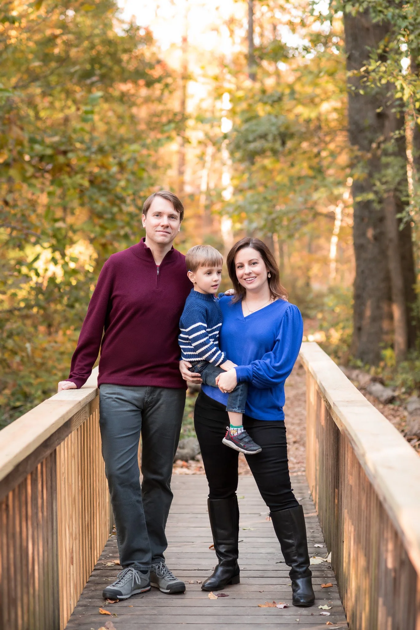 Family of three standing on a wooden bridge in a forest with autumn leaves, a father, mother, and young son smiling.