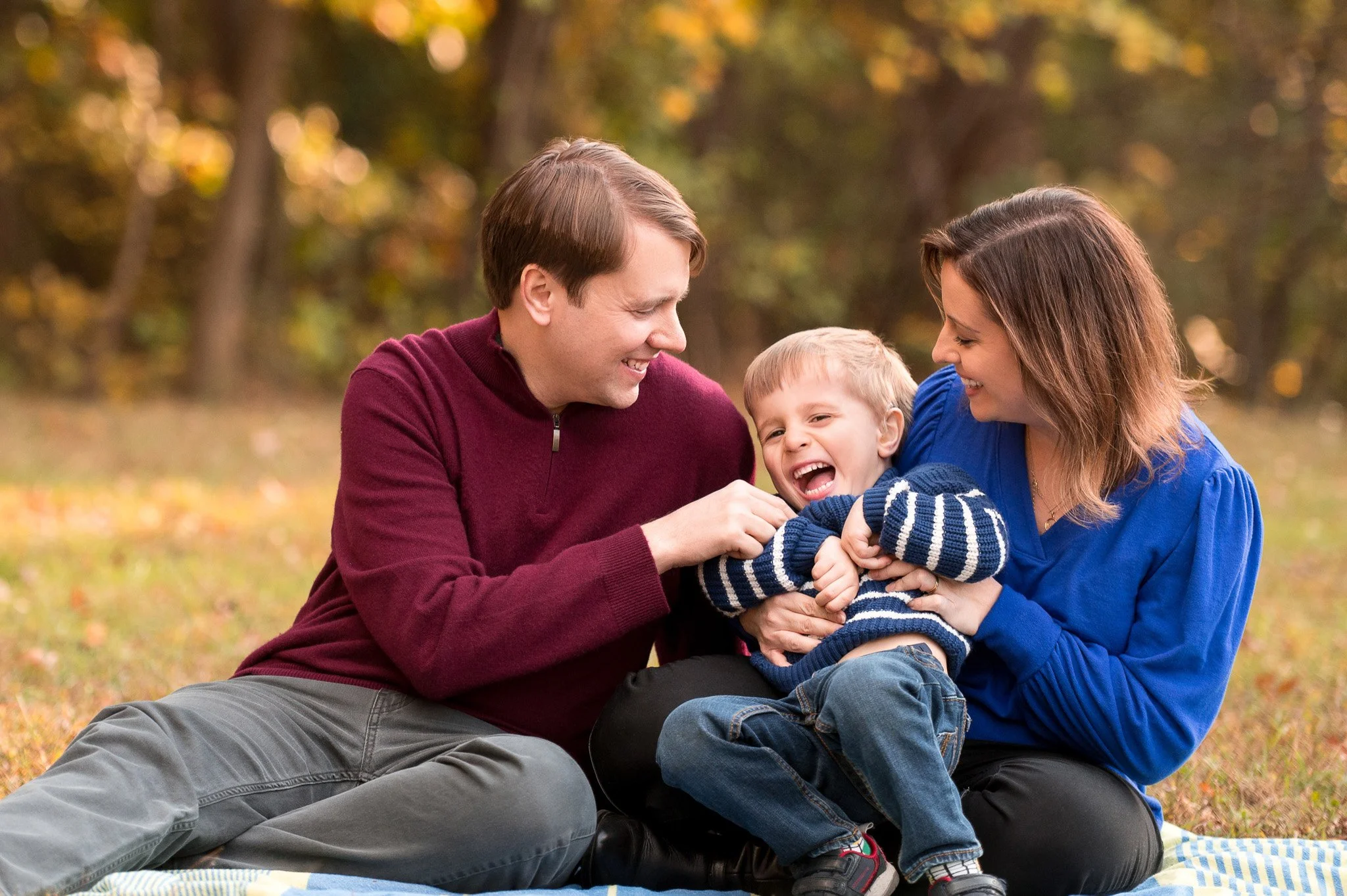 A family of three sitting on a blanket in a park, laughing and playing with their young son, surrounded by trees with autumn leaves.
