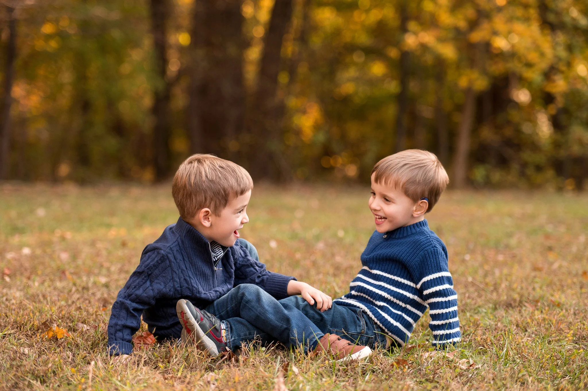 Two young boys sitting on the grass in a park, smiling and playing together with trees and autumn leaves in the background.