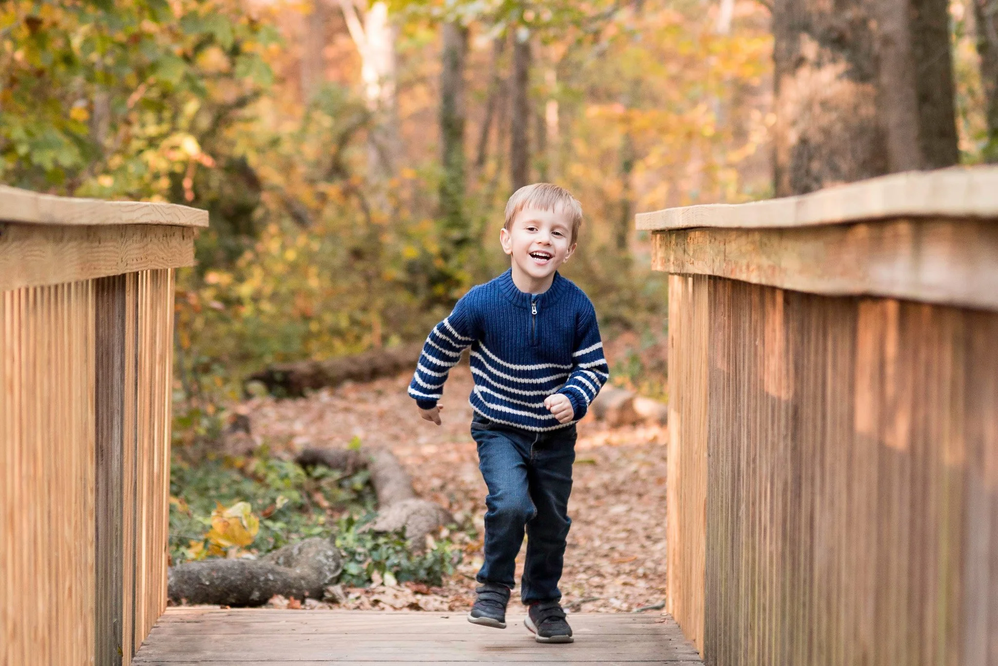 Young boy running on wooden bridge through a forest with autumn leaves and trees in the background.