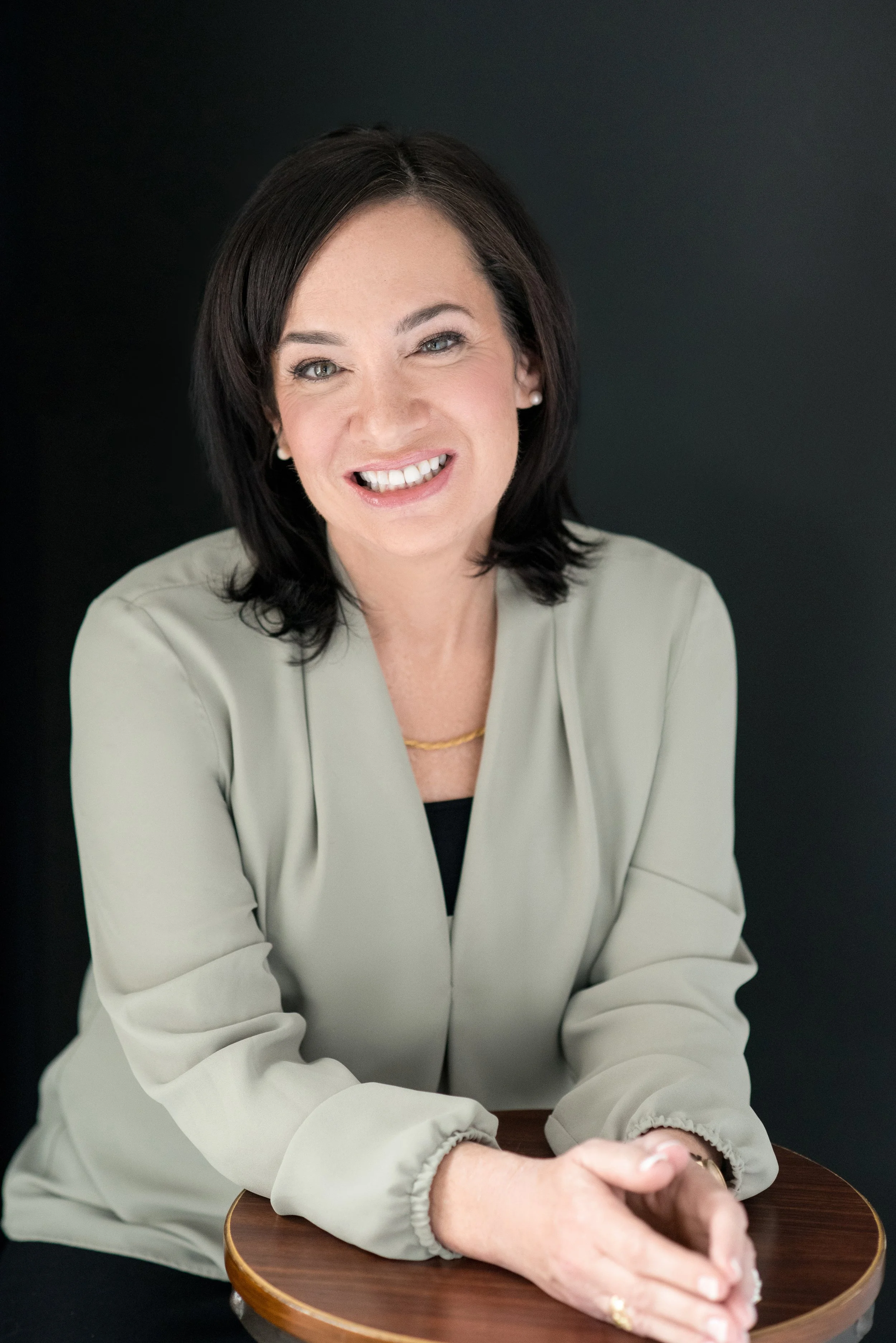 A woman with dark hair and a light-colored blouse smiling at the camera, sitting at a round wooden table against a black background.